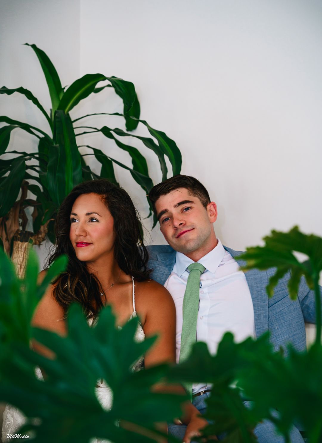 Couple poses intimately among indoor plants with green tie and formal attire during their anniversary photoshoot.