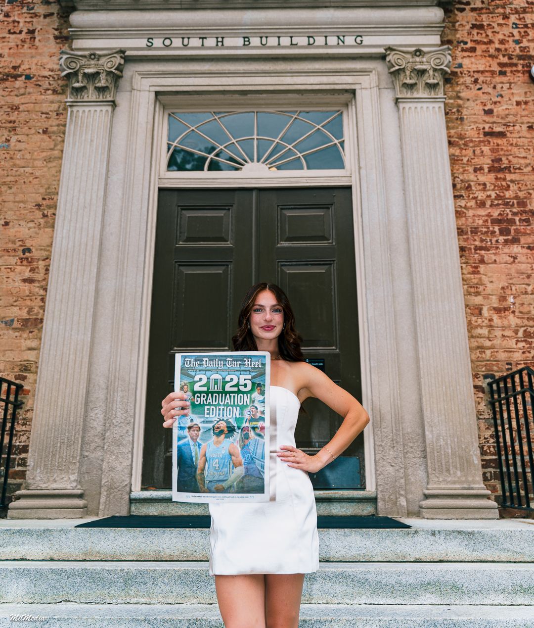 A UNC graduate poses with a campus newspaper in front of a theHistoric South Building with white columns and architectural details.