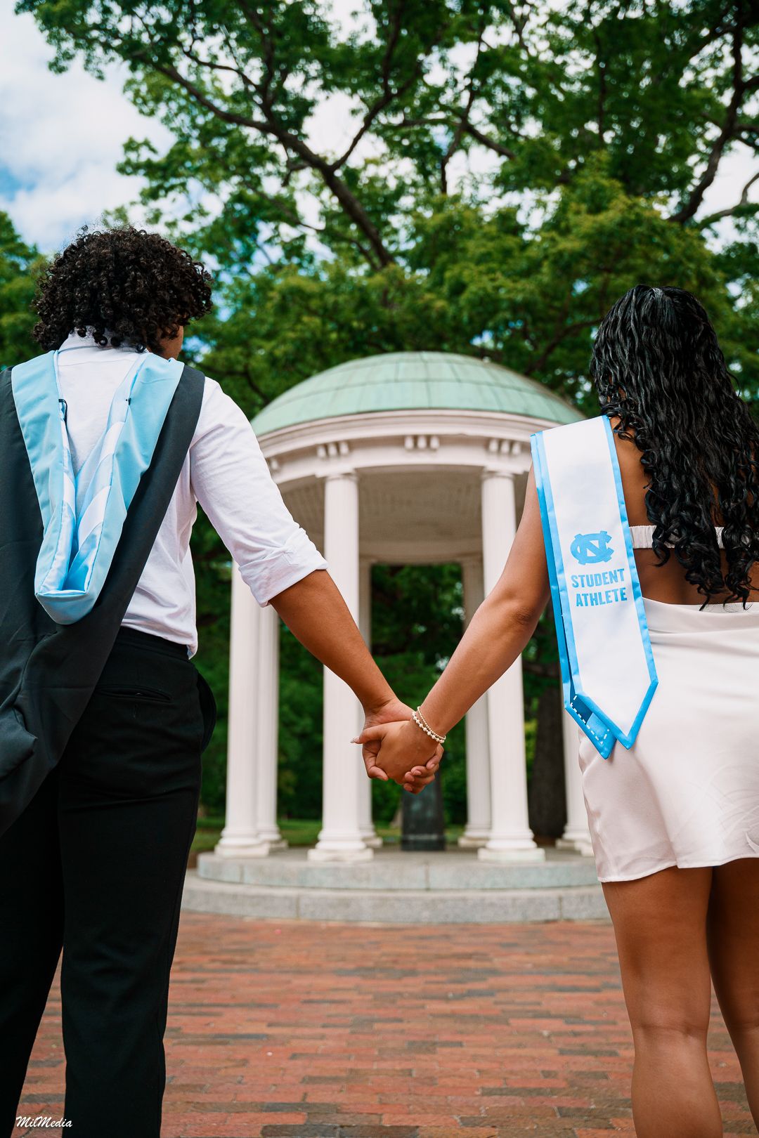 Graduates wearing blue and white sashes hold hands in front of the Old Well at UNC Chapel Hill.