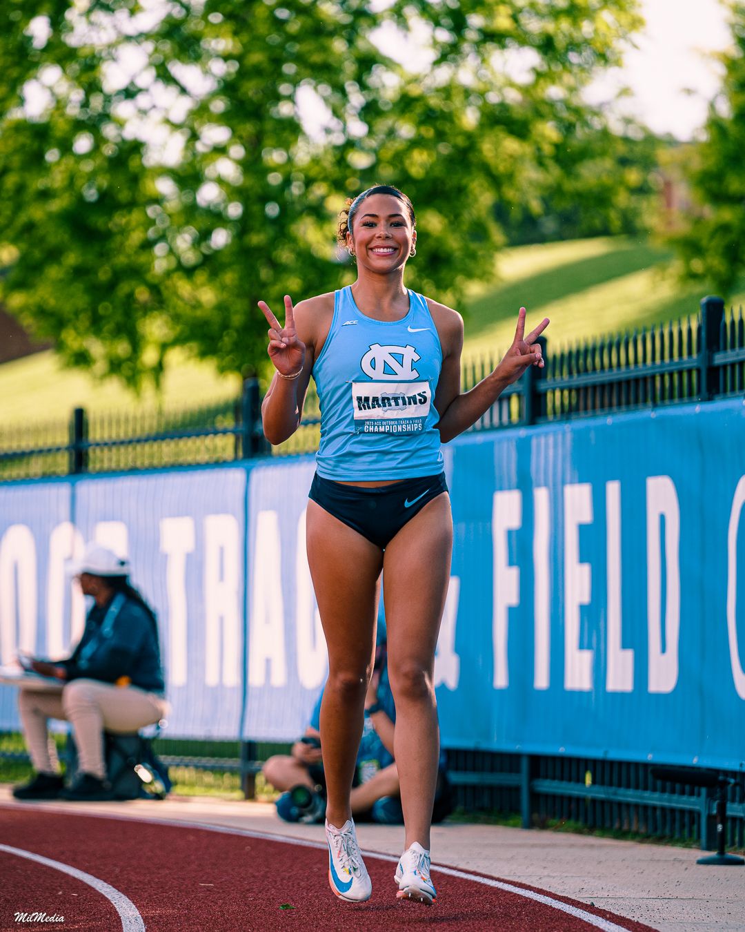 Athletes celebrate victoriously on an outdoor track with blue field signage visible in the background during the ACC Track and Field Champio