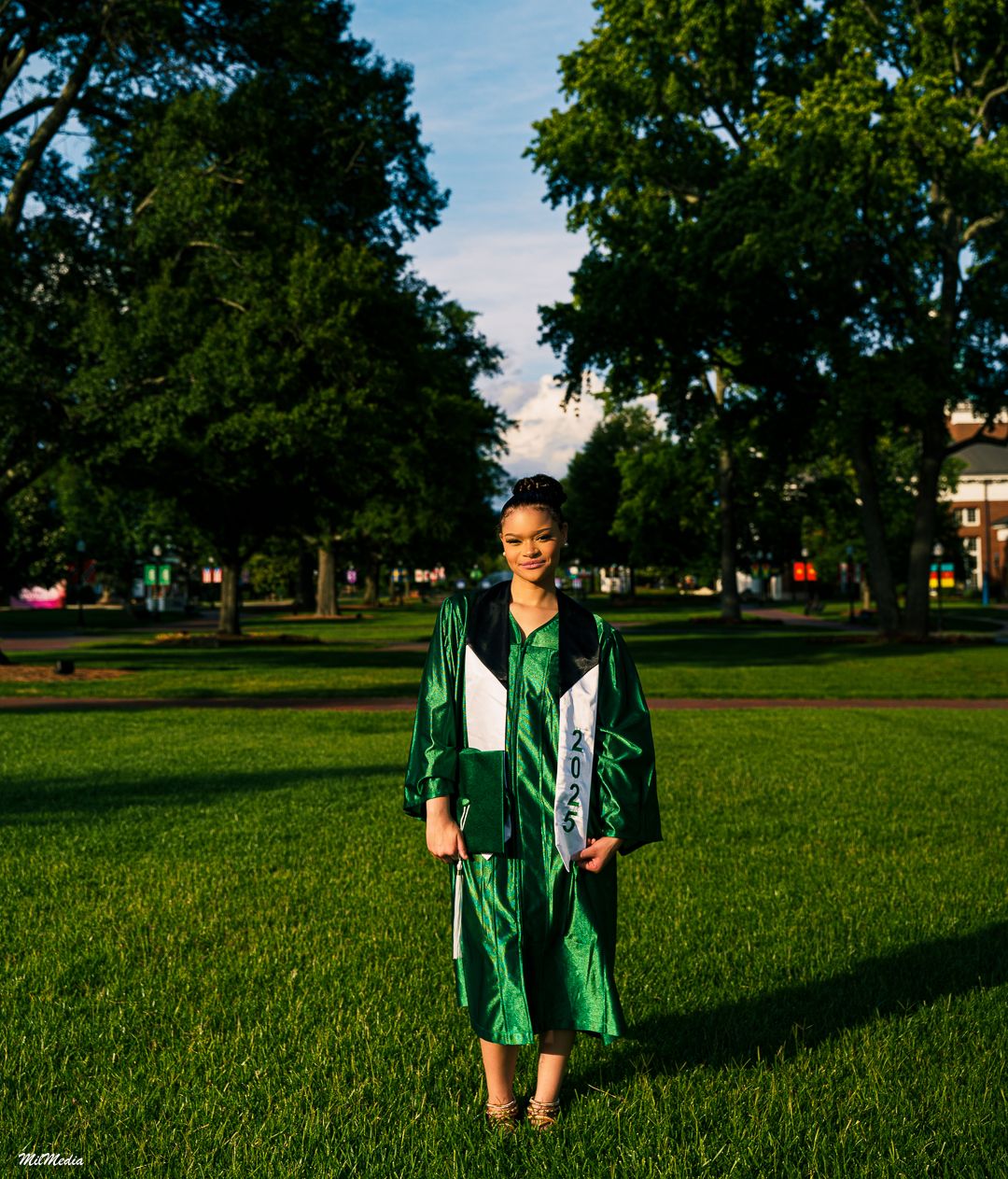 Graduate in green cap and gown stands on sunny grass field during outdoor graduation photoshoot.