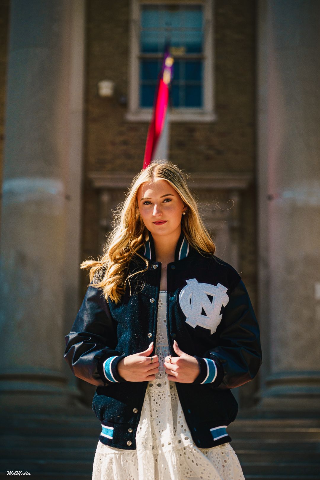 A person wearing a UNC varsity jacket and white dress poses in front of a stone building in an artistic portrait series.
