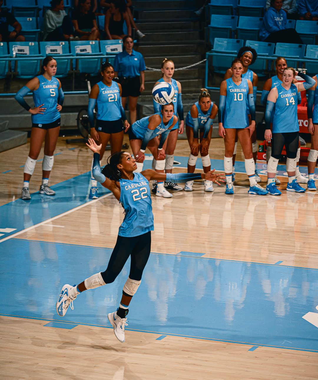 Athletes in Carolina blue uniforms practice volleyball serves on the court.