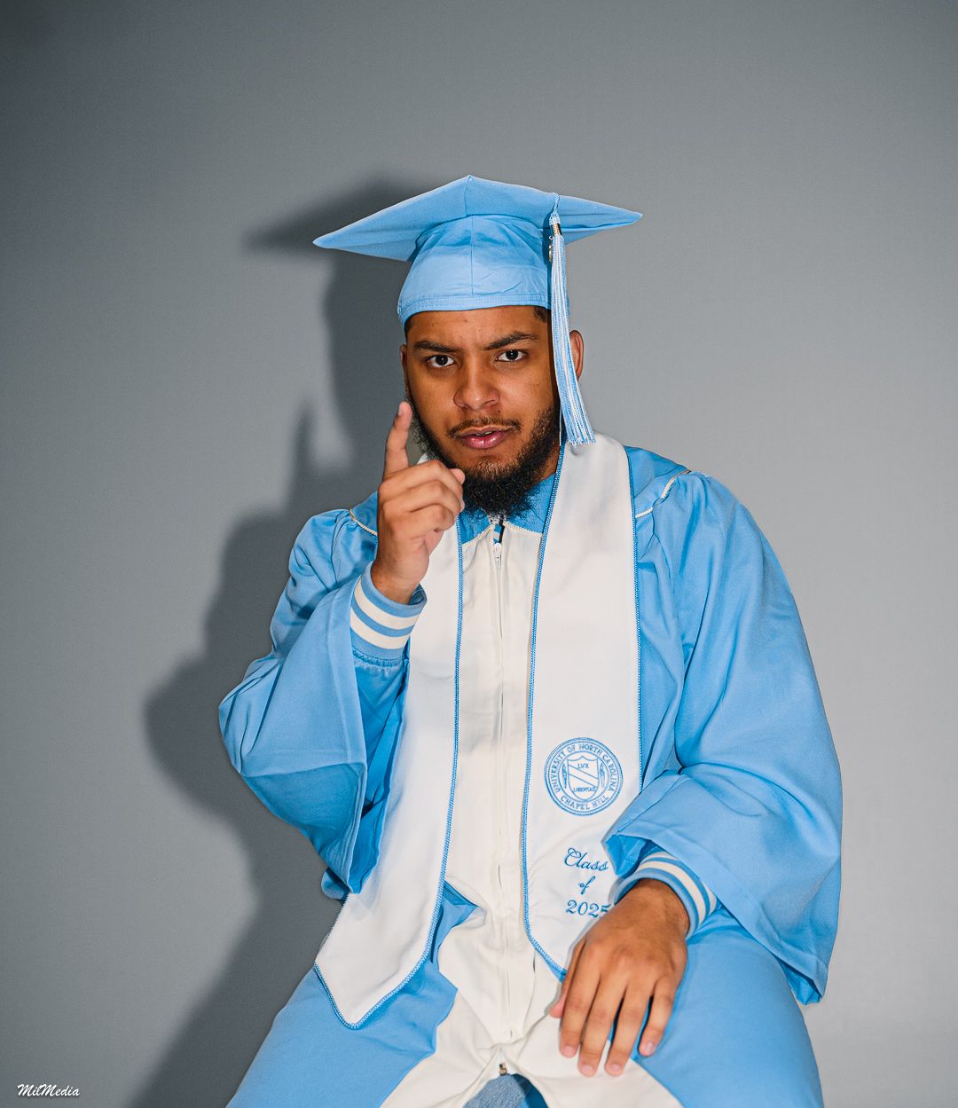 A graduate in UNC academic regalia poses casually against a grey backdrop.