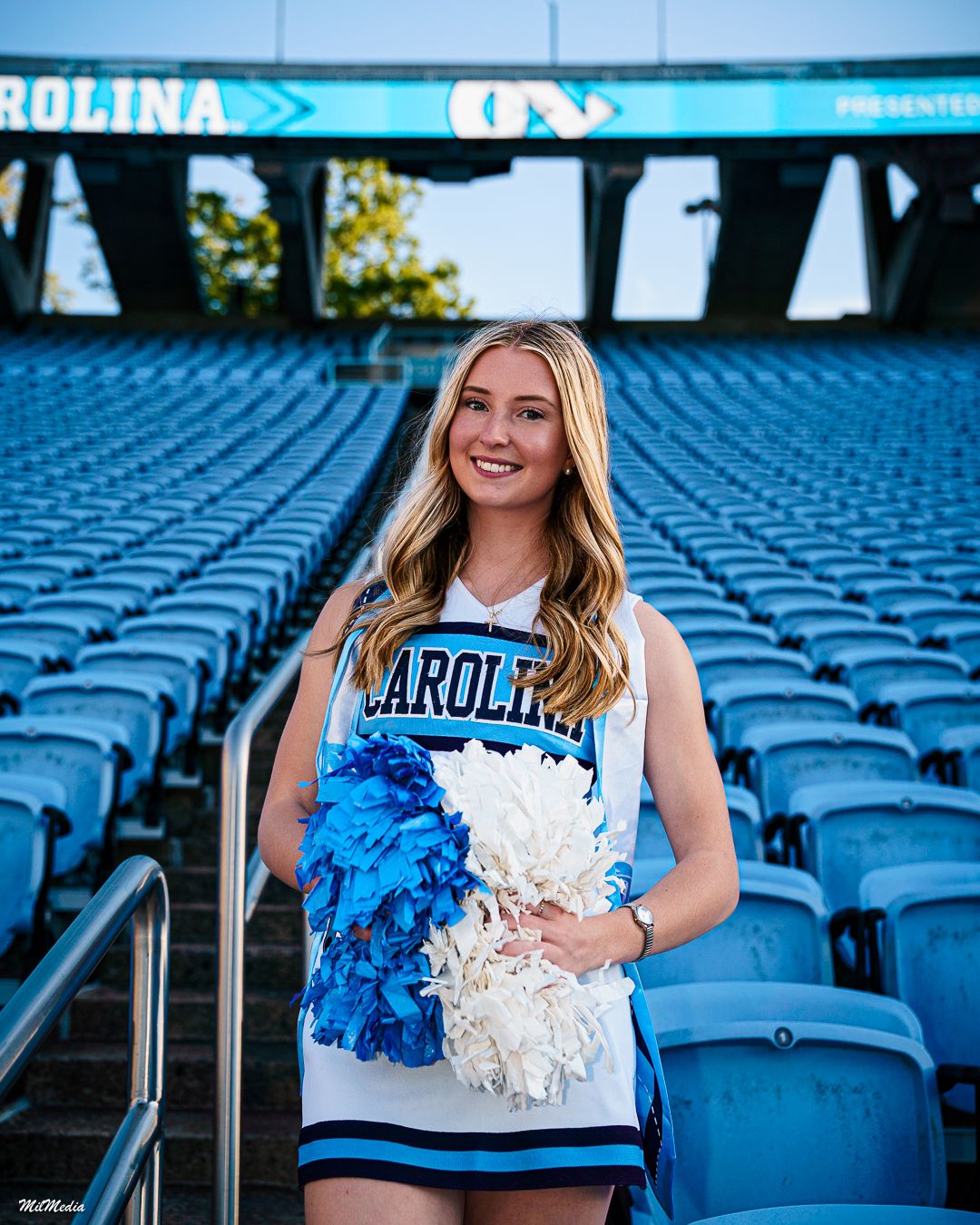 Cheerleader poses in stadium seats wearing blue and white uniform with pom poms at UNC sports venue.