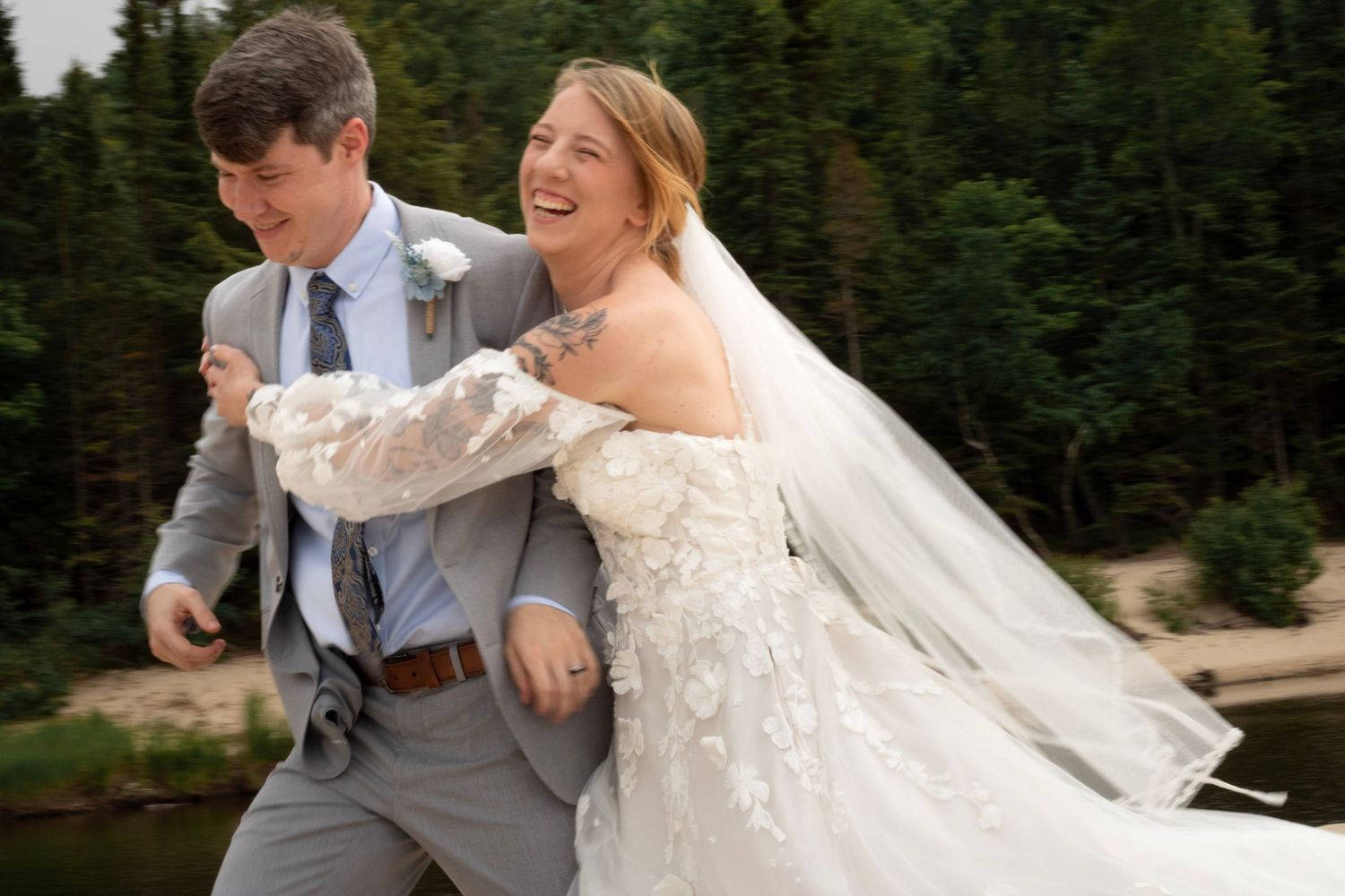 Laughing bride in lace gown and groom in gray suit share a joyful moment outdoors near a lake and pine trees.