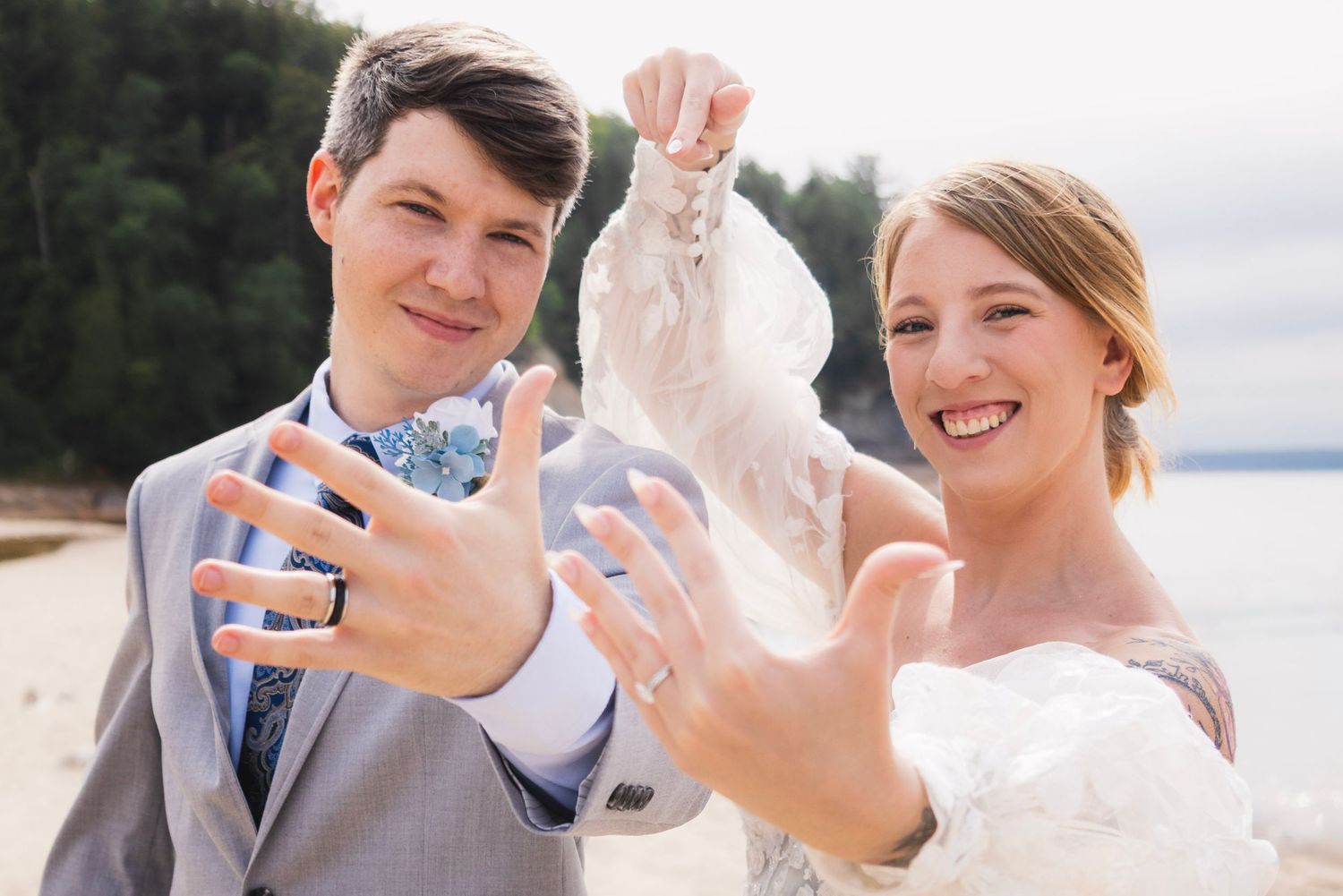 Newlywed couple showing off wedding rings on beach, groom in gray suit with bow tie, bride in white dress smiling.