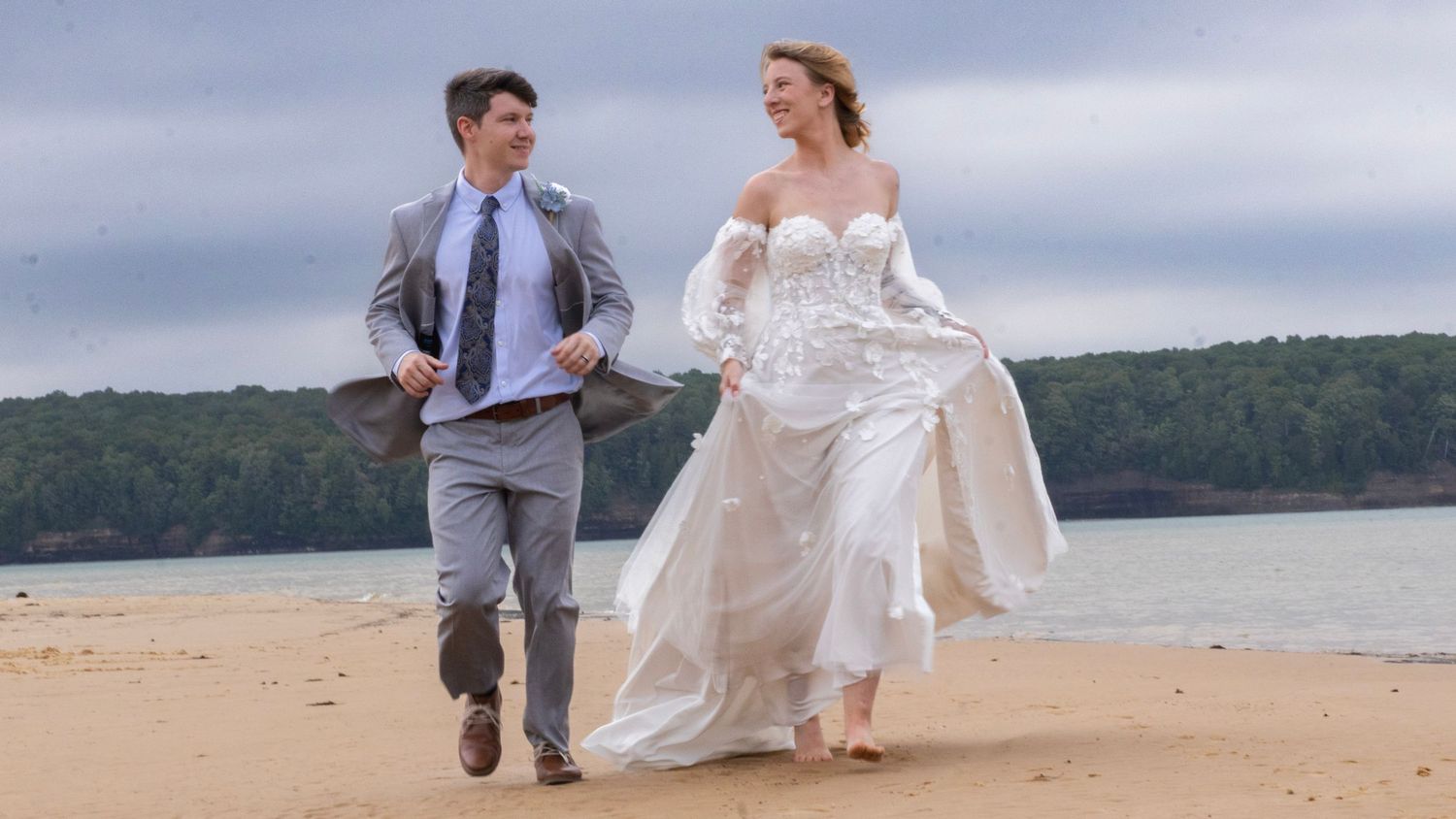 A joyful couple runs on a sandy beach, bride in white gown and groom in gray suit, with trees in the background.