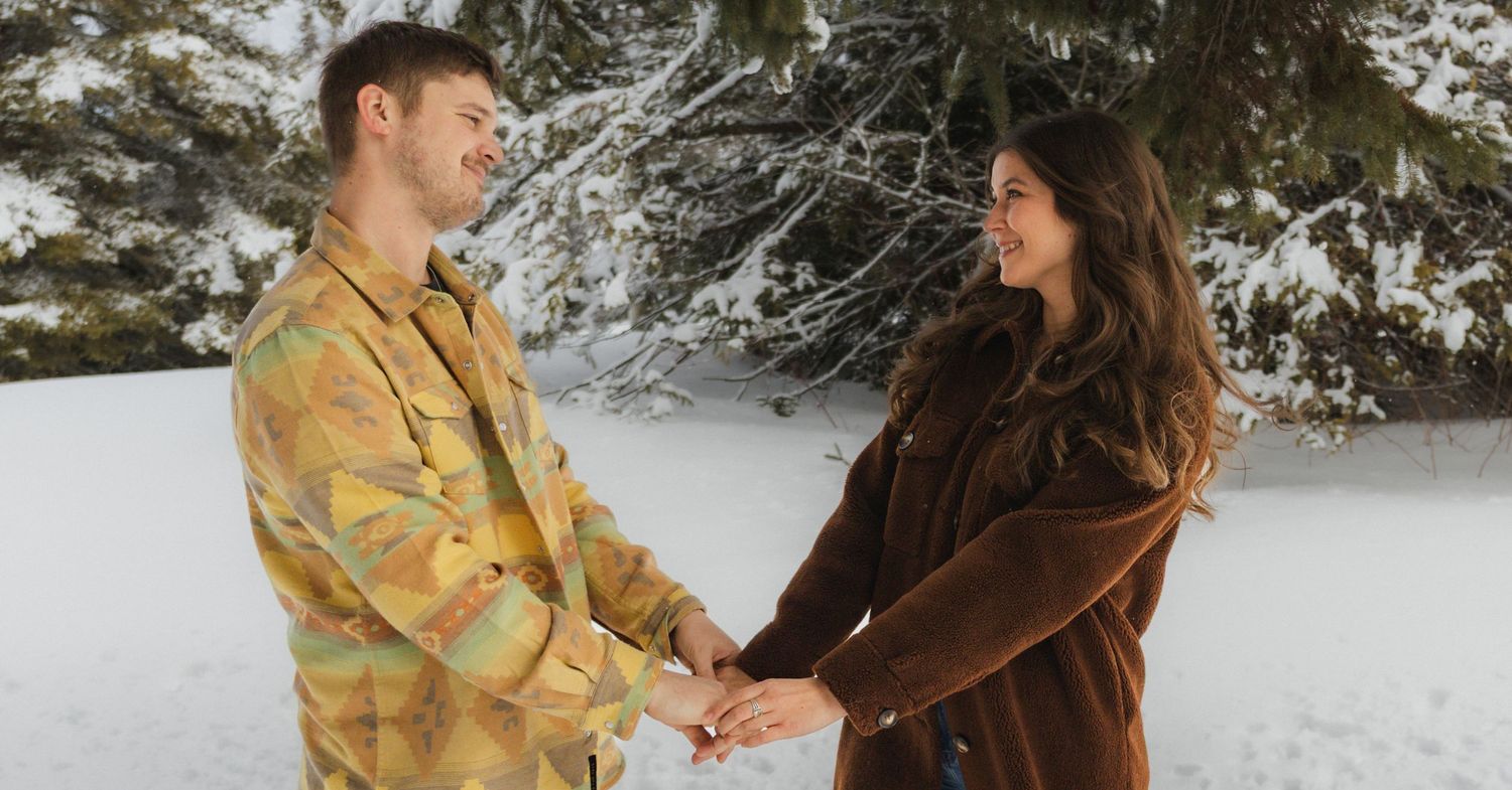 A couple stands together in a snowy winter forest wearing warm coats and enjoying a romantic moment.