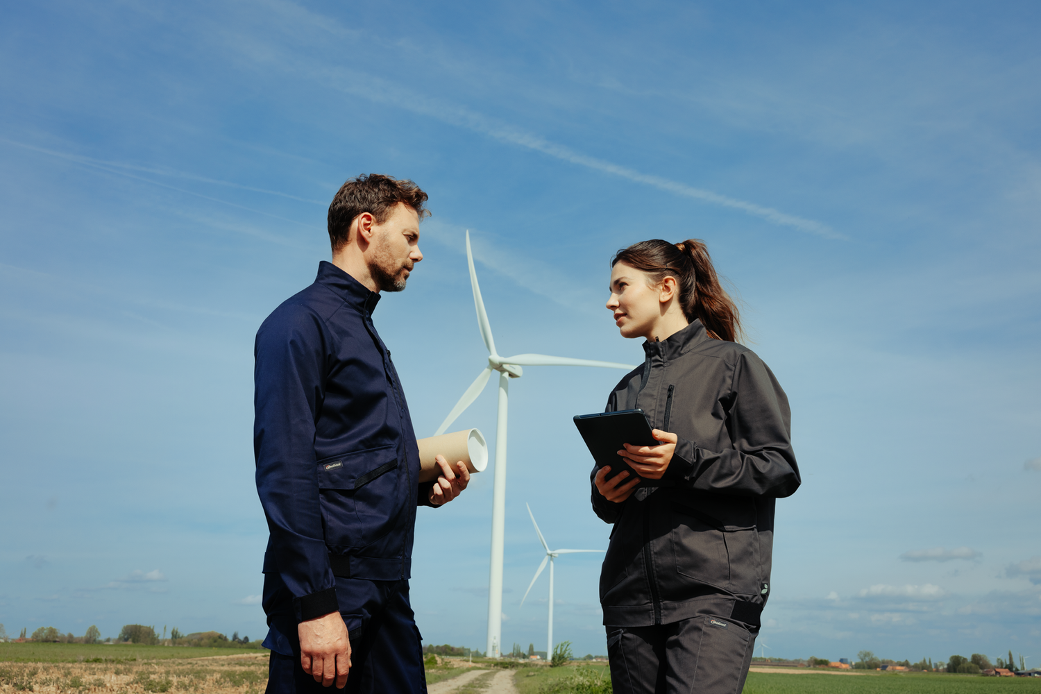 Un homme qui discute avec une femme qui prend des notes devant un champs d’éolienne