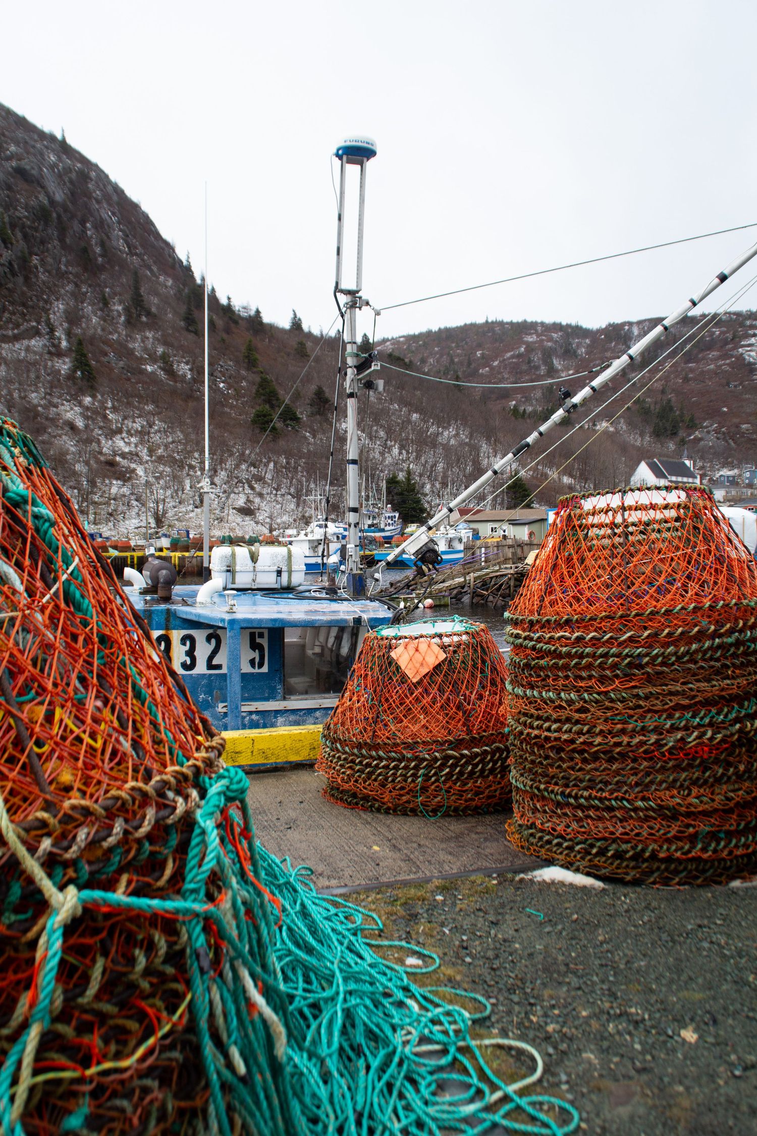 Pièges à pêche oranges et corde turquoise avec en arrière plan un bateau de pêche bleu et une montagne de pins.