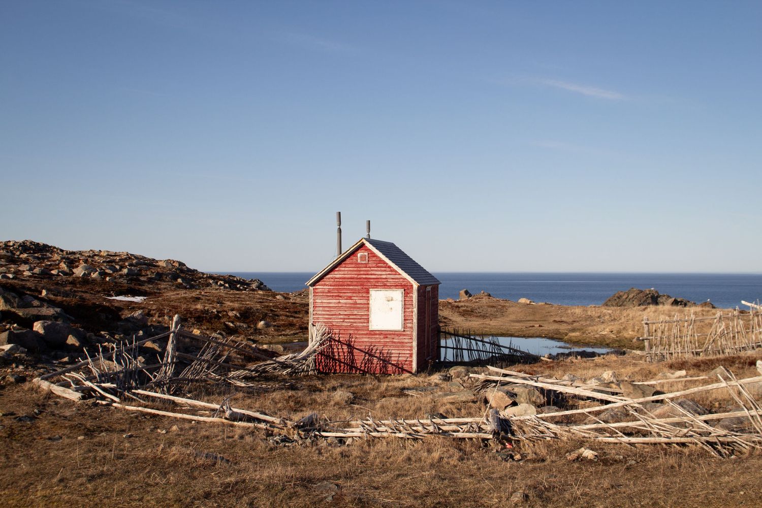 Cabane rouge et blanche à Bonavista entourée d’une vieille clotûre au sol avec la mer an arrière plan.