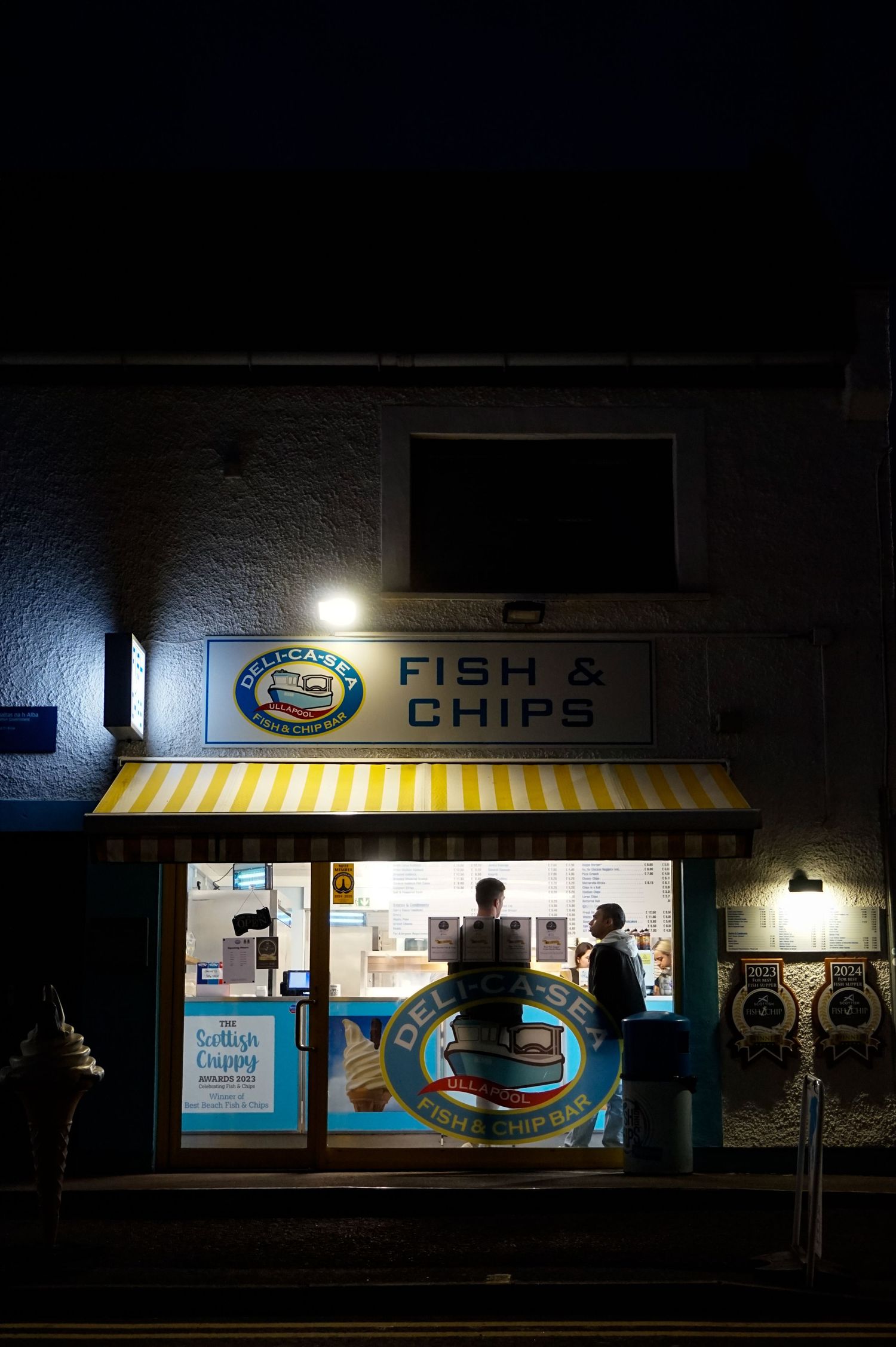 Photo de nuit d’une devanture éclairée d’une boutique de fish&chips jaune avec deux hommes à l’intérieur.