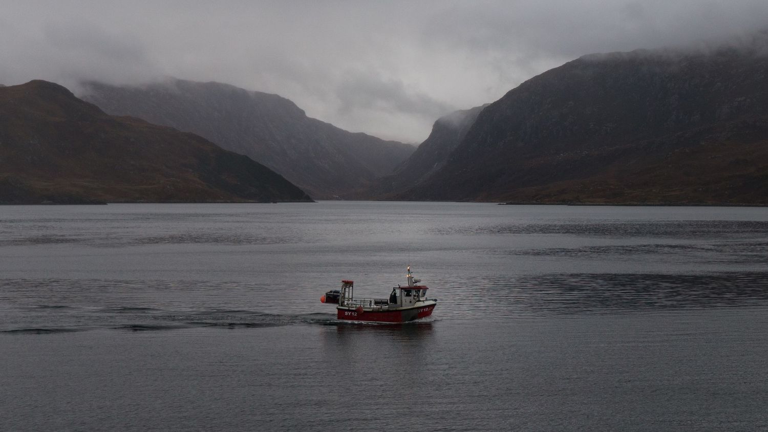 Bateau de pêche écossais rouge et blanc au milieu de la mer avec des montagnes embrumées en arrière plan.