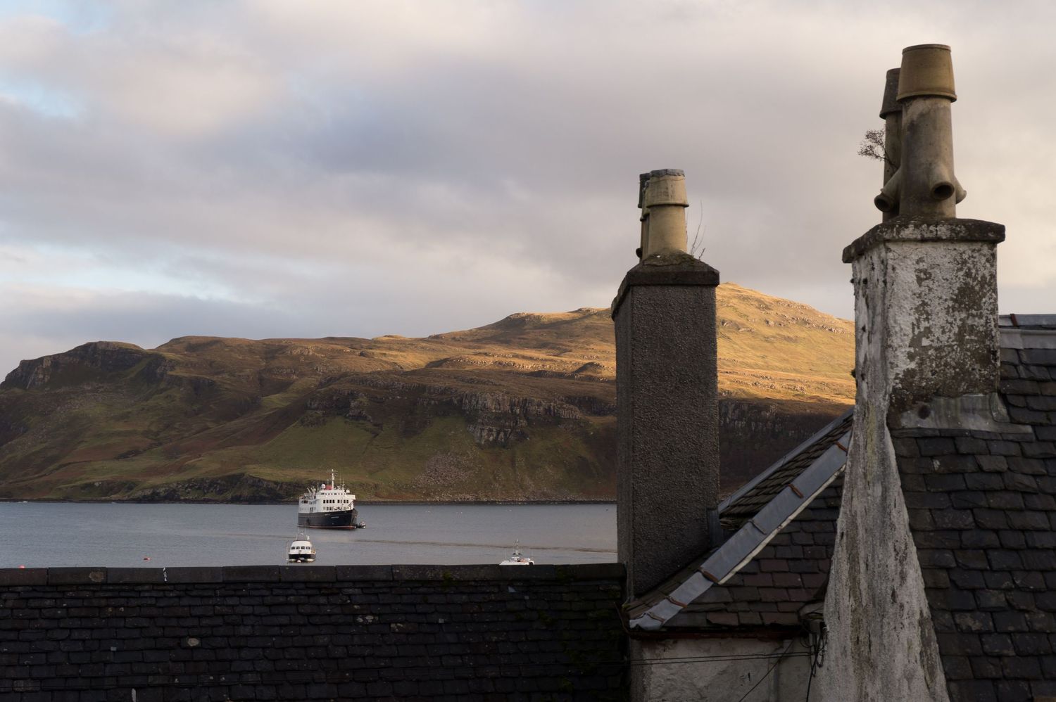 Toit d’une maison de Portree en Ecosse avec en arrière plan des bateaux sur le mer et une montagne.