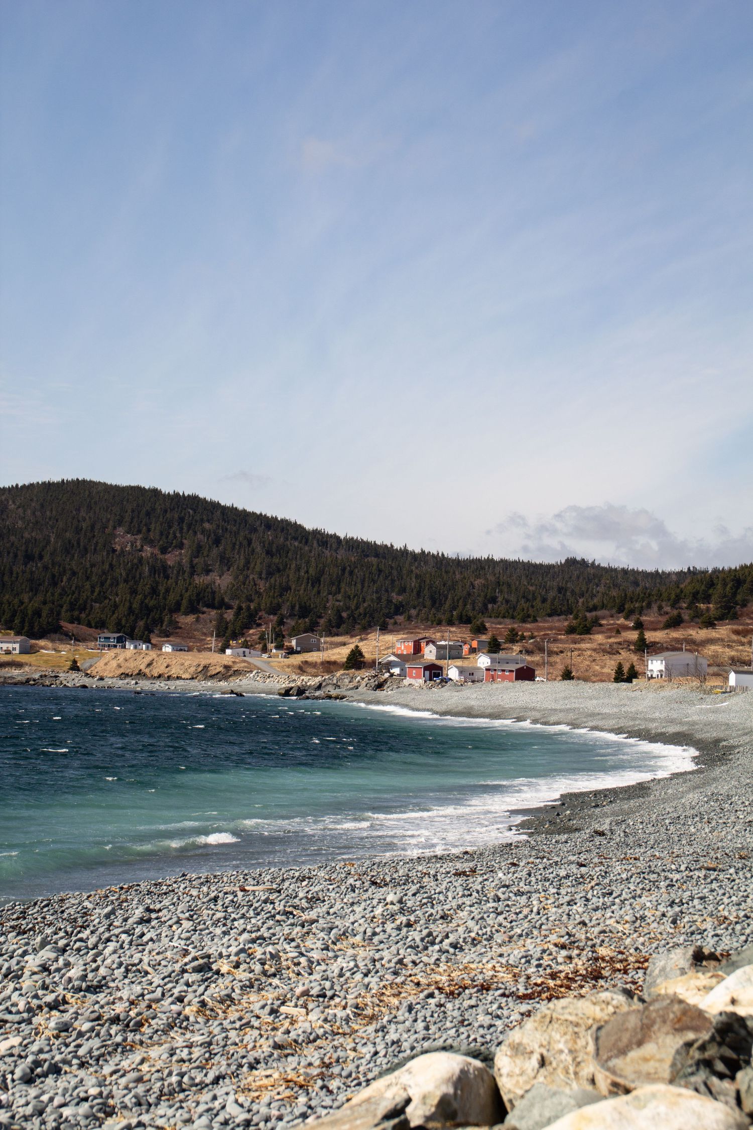 Plage de galets à Terre-Neuve avec une eau turquoise et bleu profond avec en arrière plan maisons et montage recouverte de pins.
