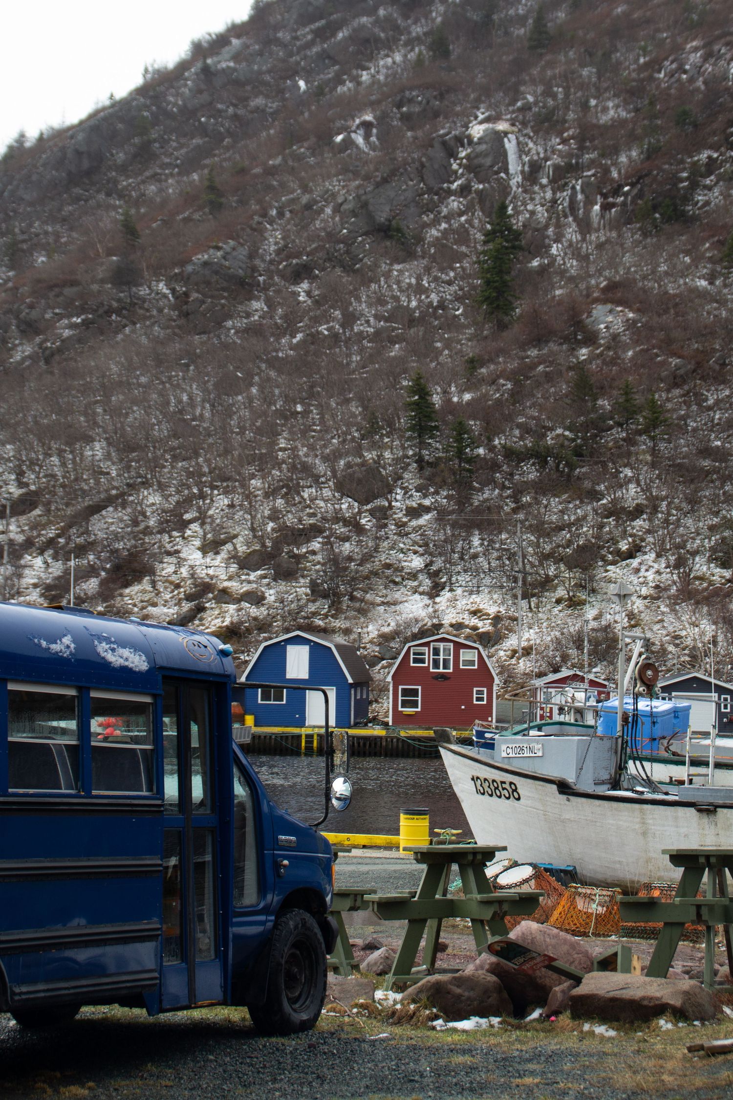 Bus d’école bleu avec en arrière plan le port de pêche, un bateau blanc et deux hangars, un bleu et un rouge.