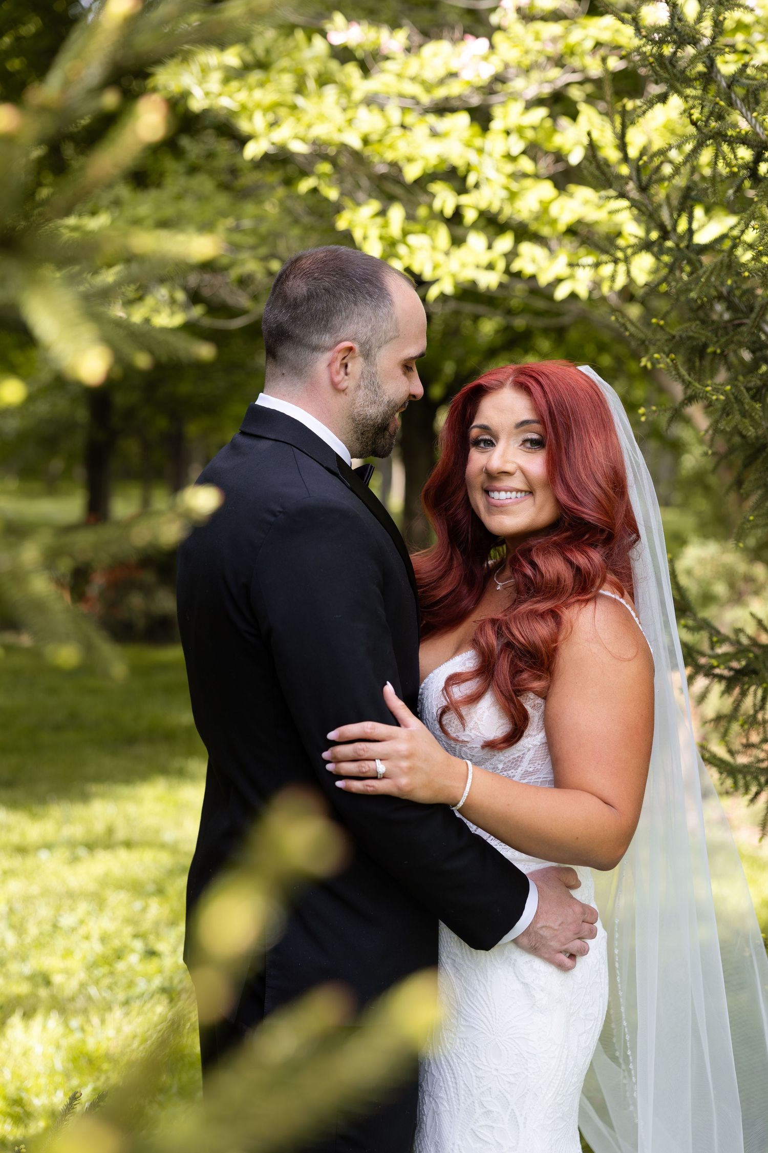 Bride with red hair in white gown and veil smiles at groom in black tuxedo, embracing outdoors in lush green garden.