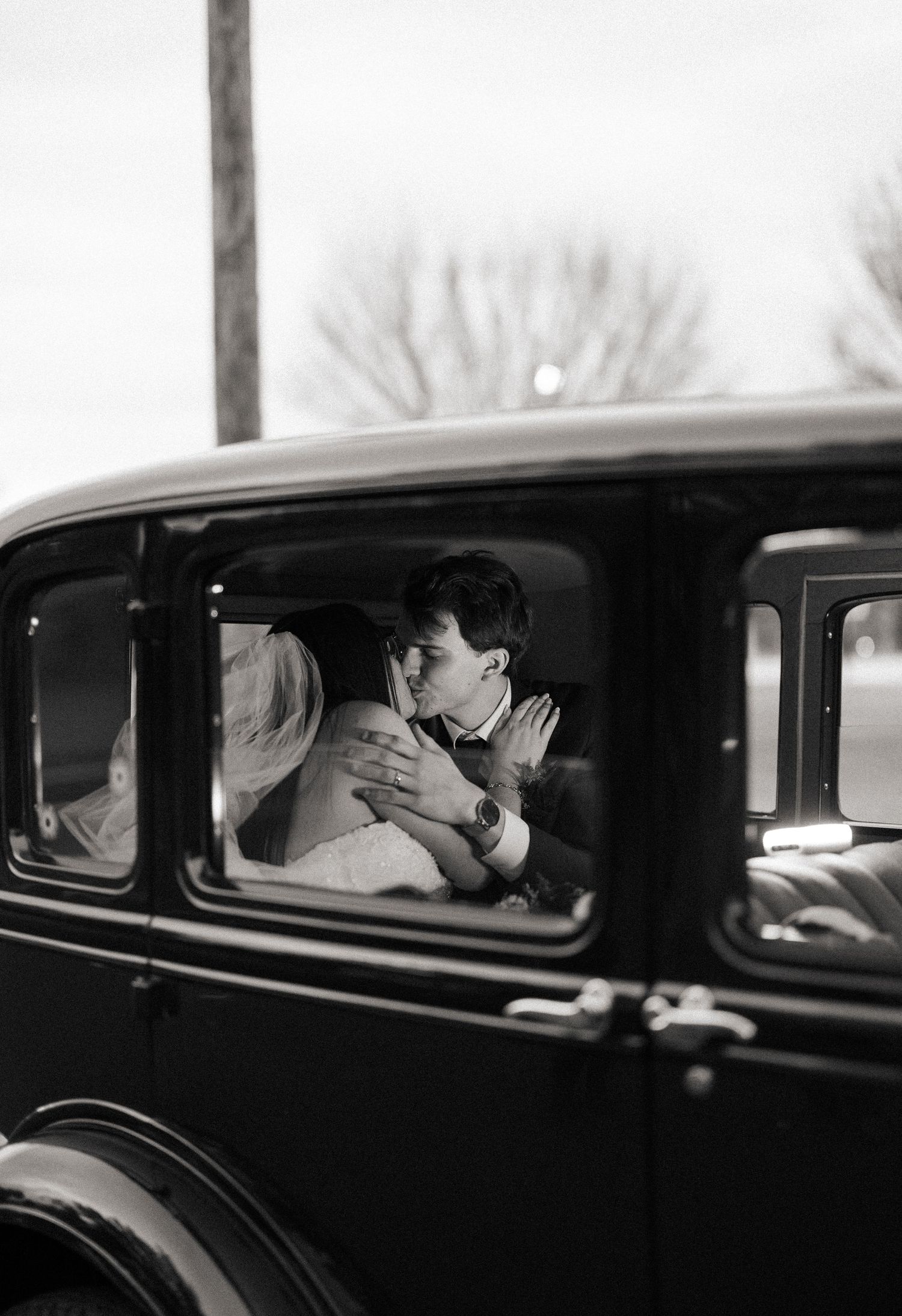 Black and white photo of a couple sharing a romantic kiss inside a vintage classic car, captured through the window.