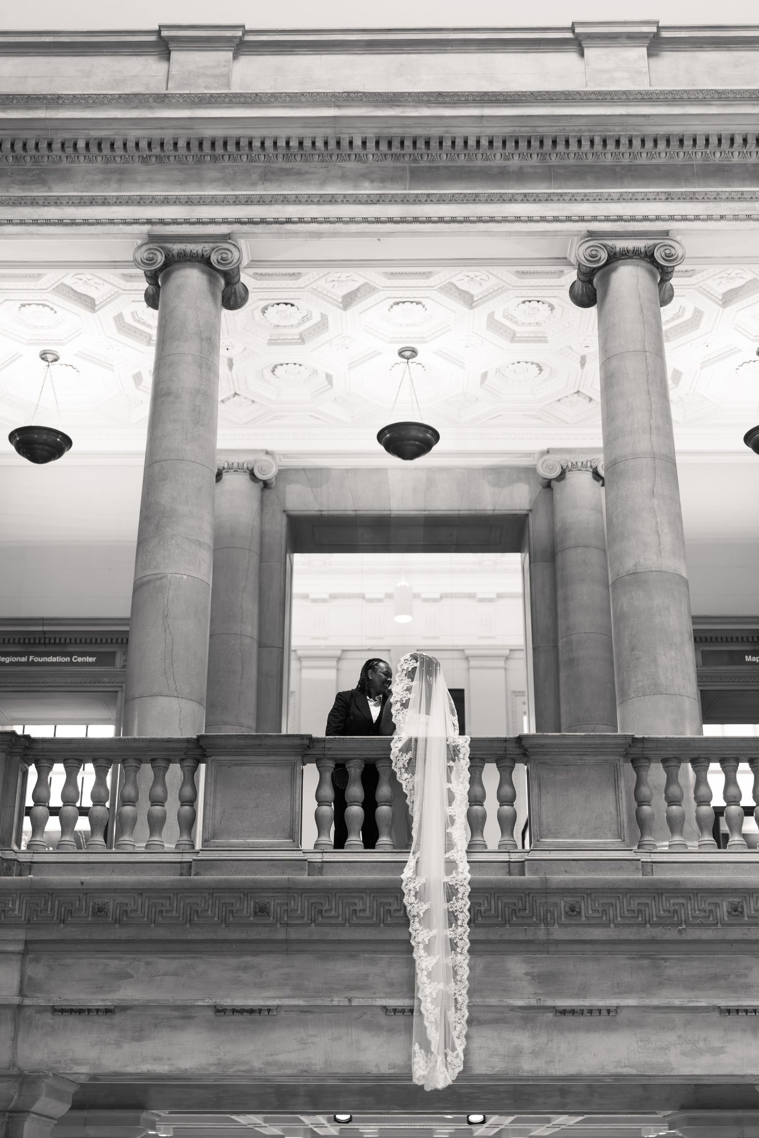 Black and white wedding photo of bride and groom on grand balcony with tall columns in elegant historic building.