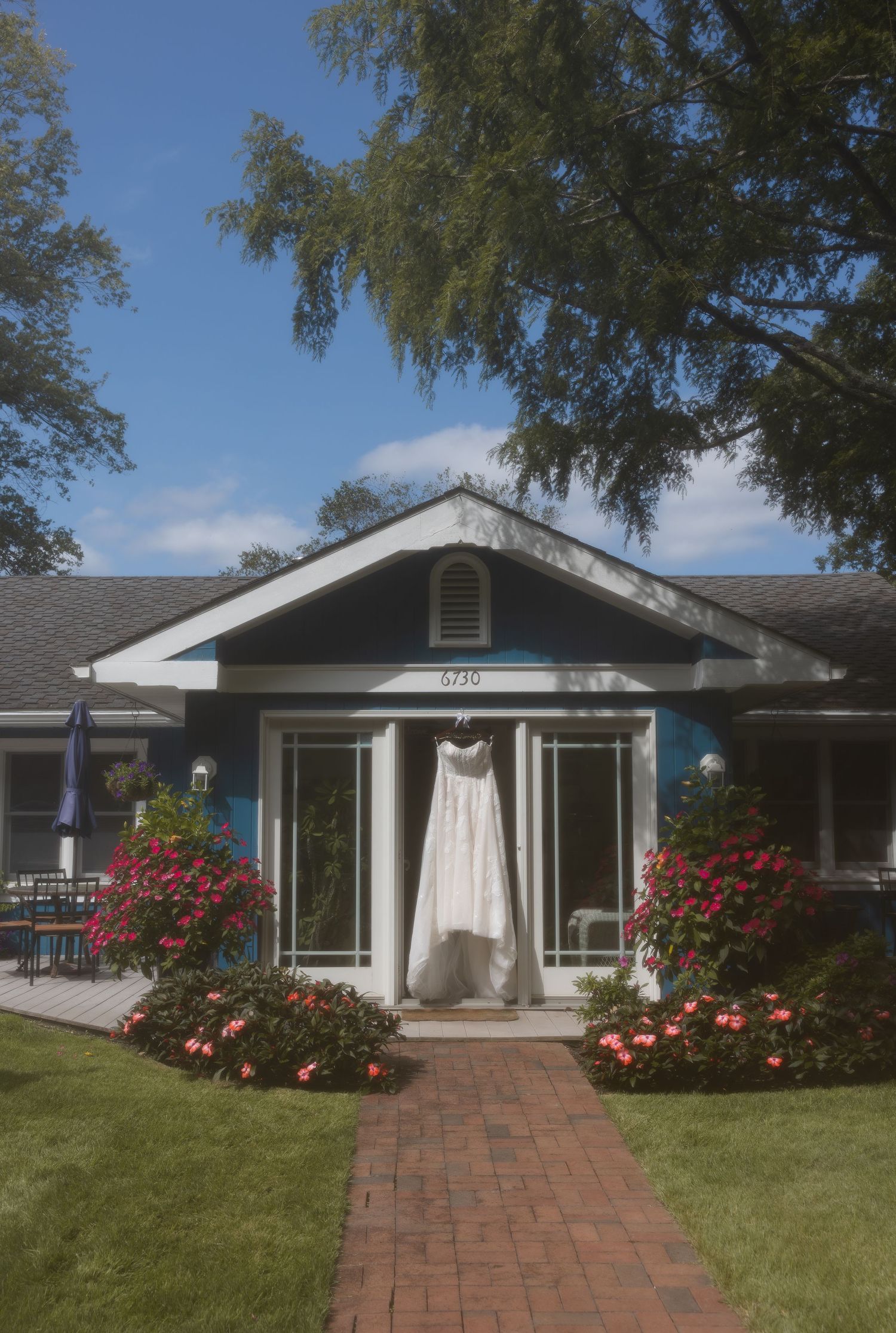 White wedding dress hanging outside a blue craftsman house with brick pathway and blooming flowers under blue sky.