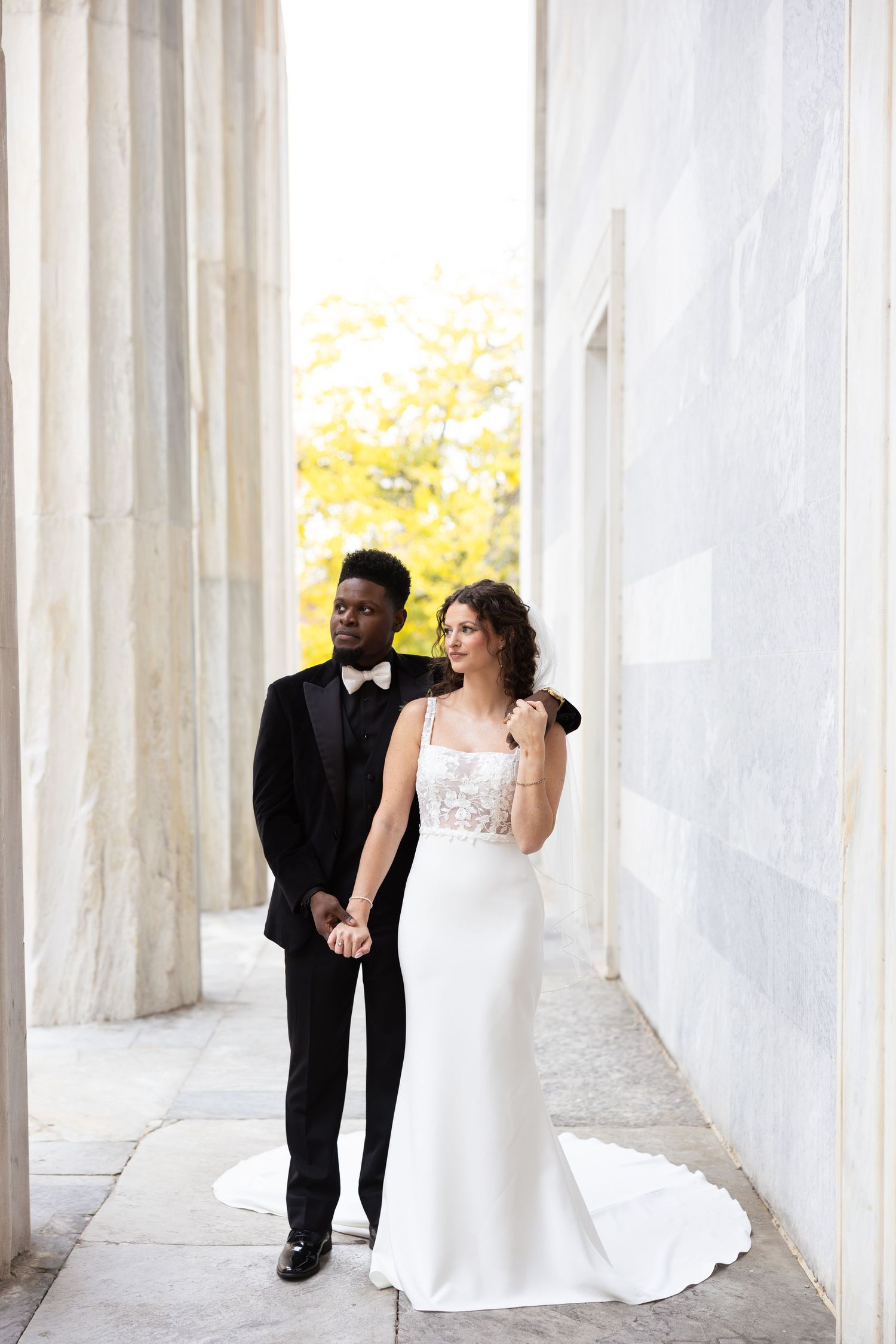 Wedding couple poses between white marble columns in an architectural setting.