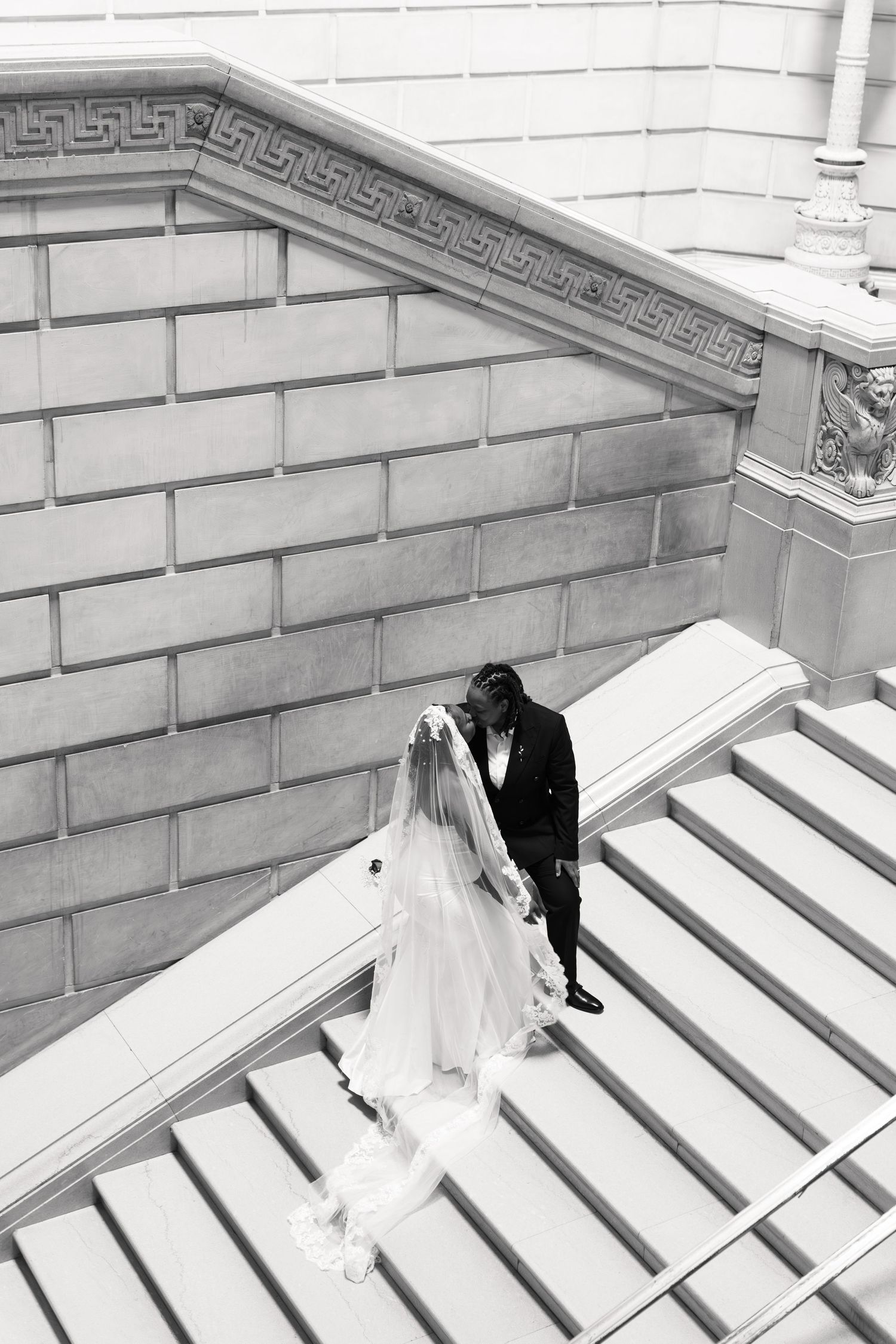 Black and white photo of bride and groom walking down grand staircase, wedding veil trailing behind.