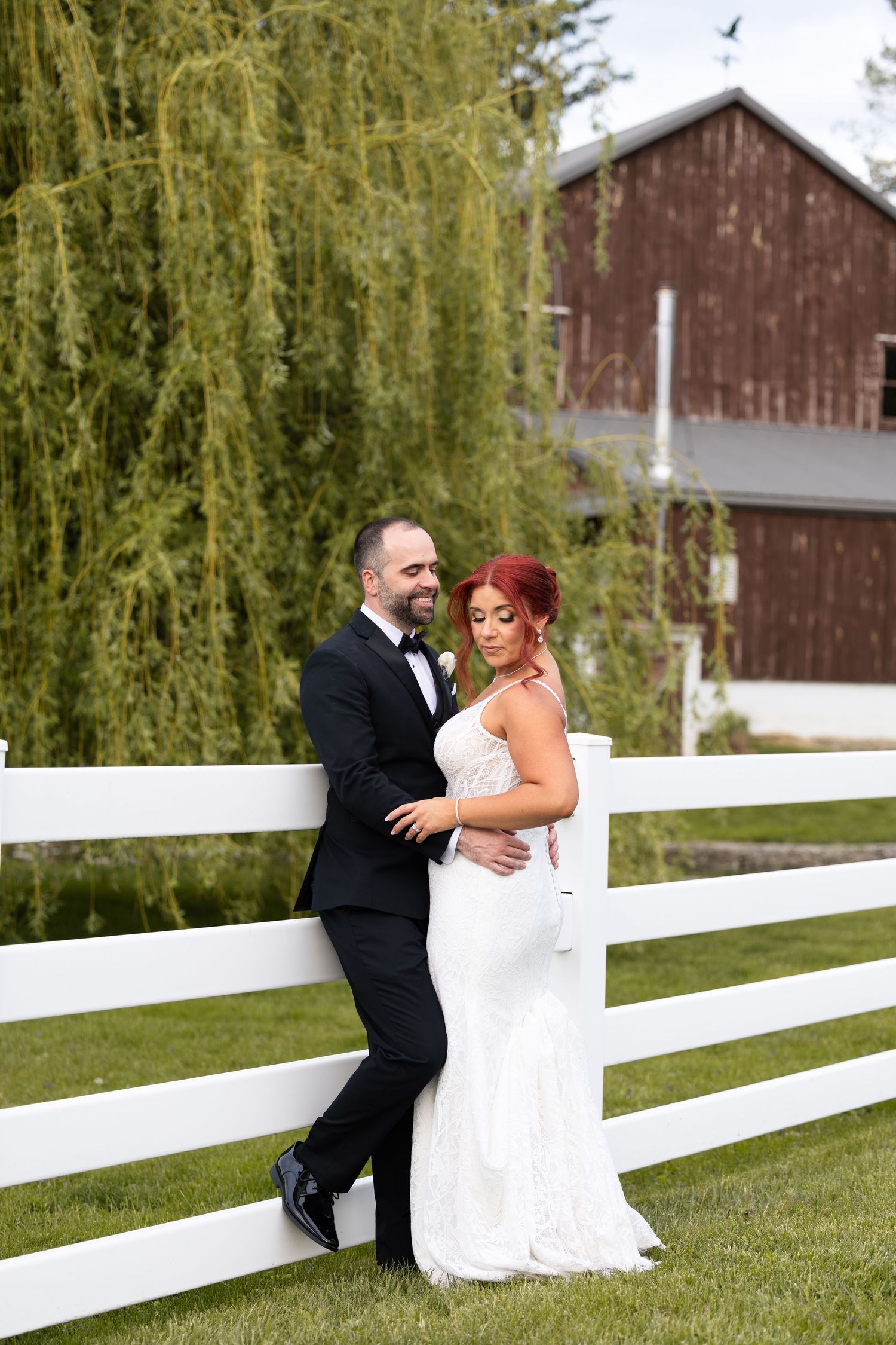 Bride and groom pose by white fence with red barn and willow tree backdrop on their wedding day.
