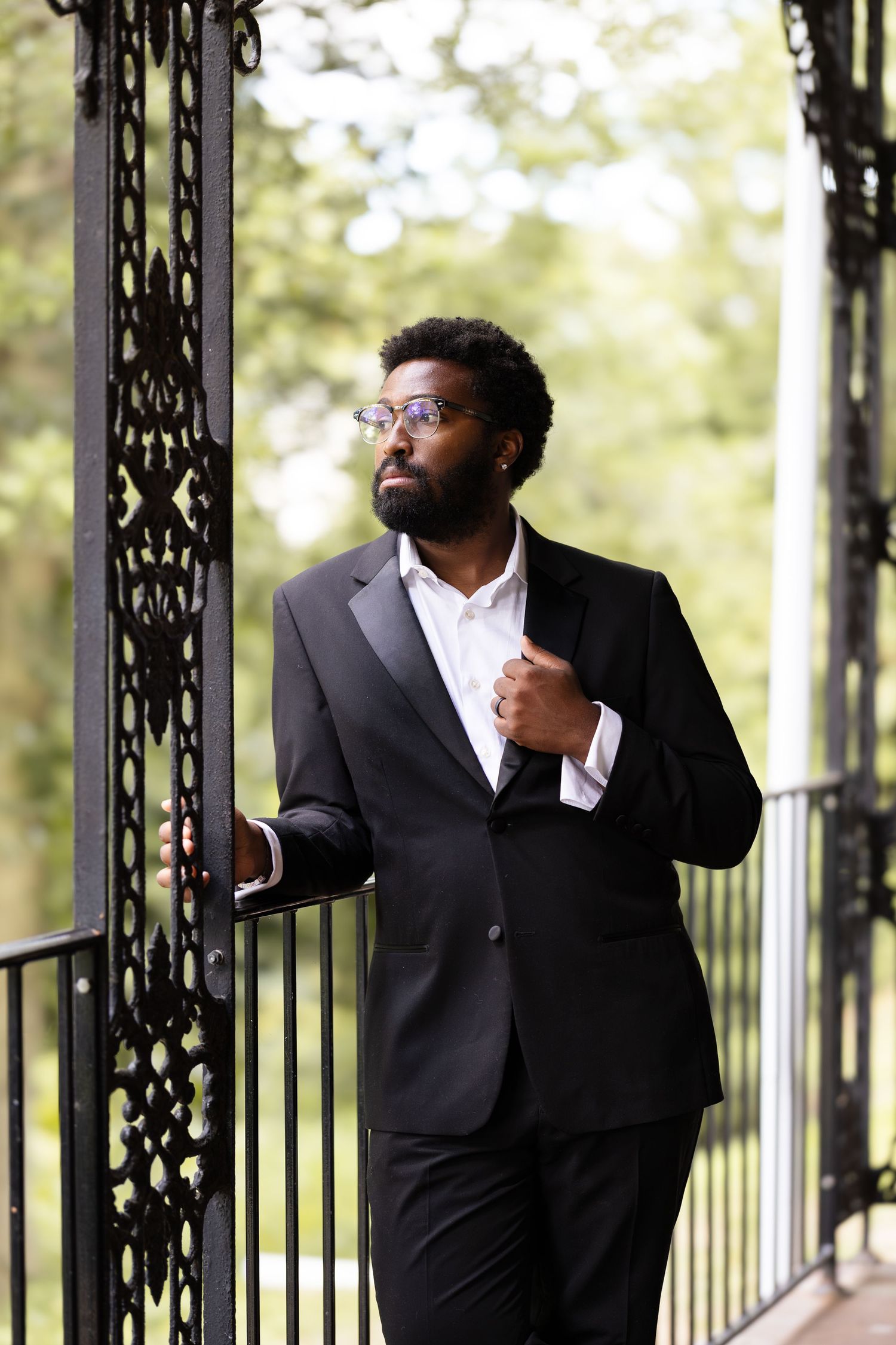 A well-dressed Black man in a black suit and glasses stands thoughtfully by ornate iron railings outdoors.