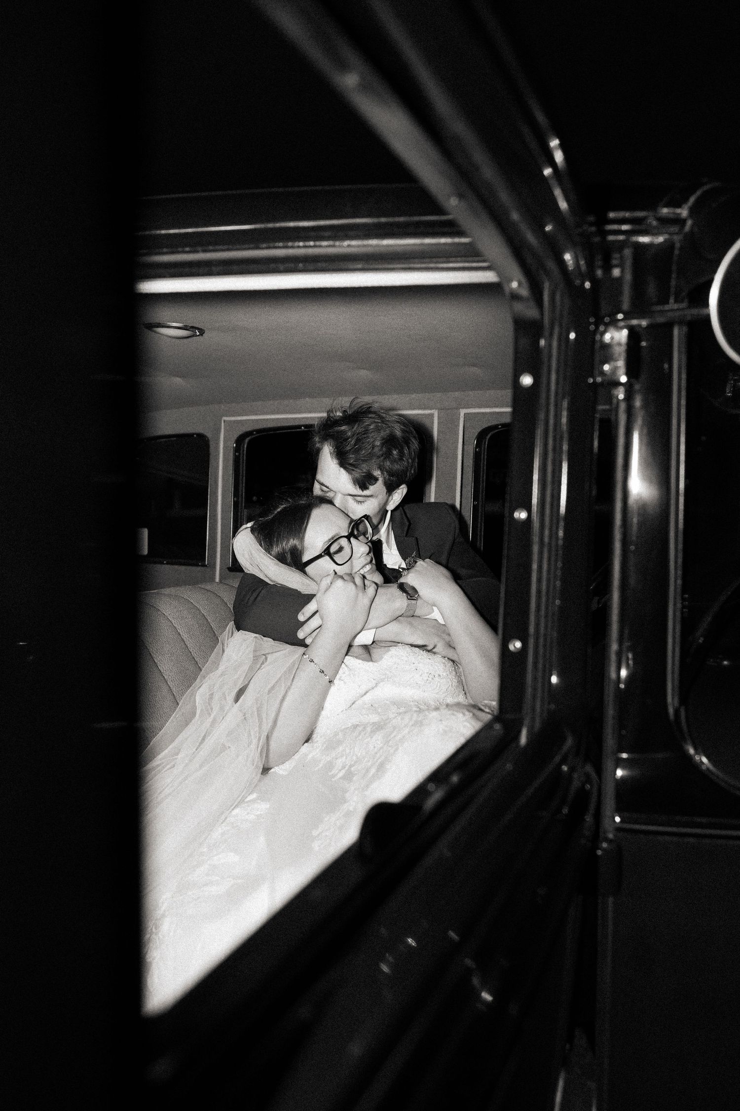 Black and white photo of a couple sharing a romantic kiss inside a vintage limousine on their wedding day.