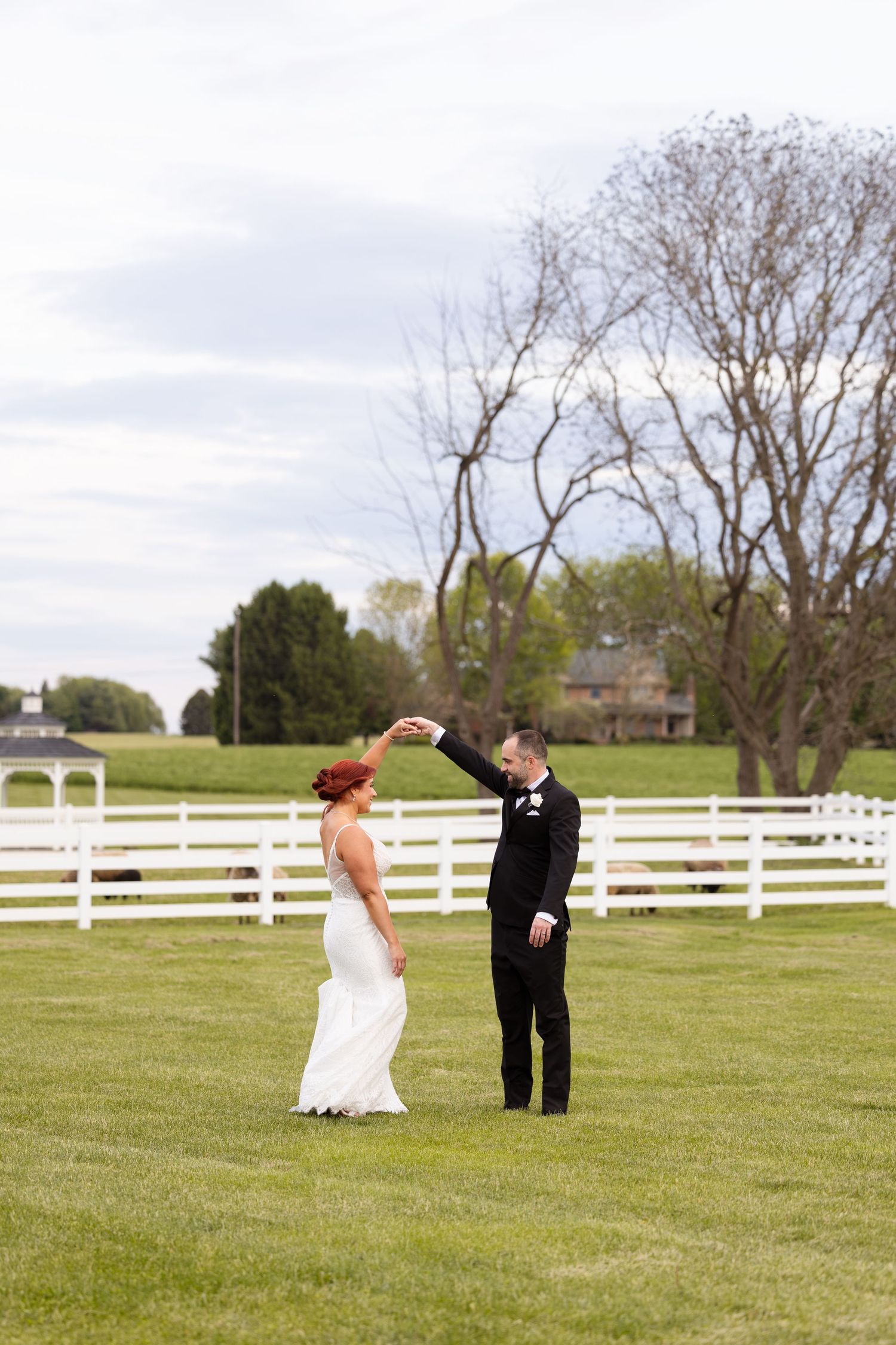 Bride and groom dancing outdoors on green lawn with white fence and trees in background at elegant wedding venue.