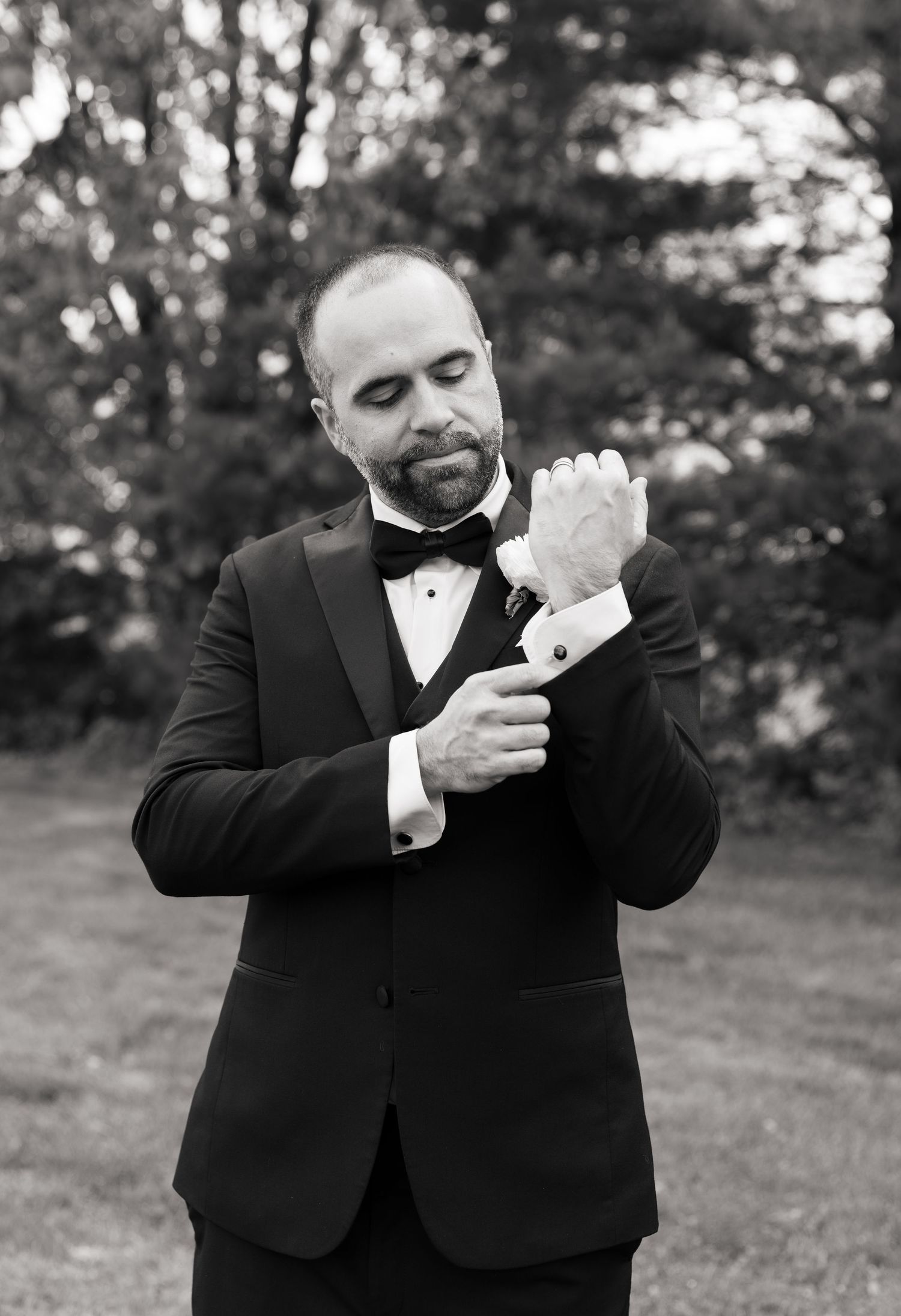 A groom in a tuxedo and bow tie adjusts his cufflinks outdoors in a black and white wedding photo.
