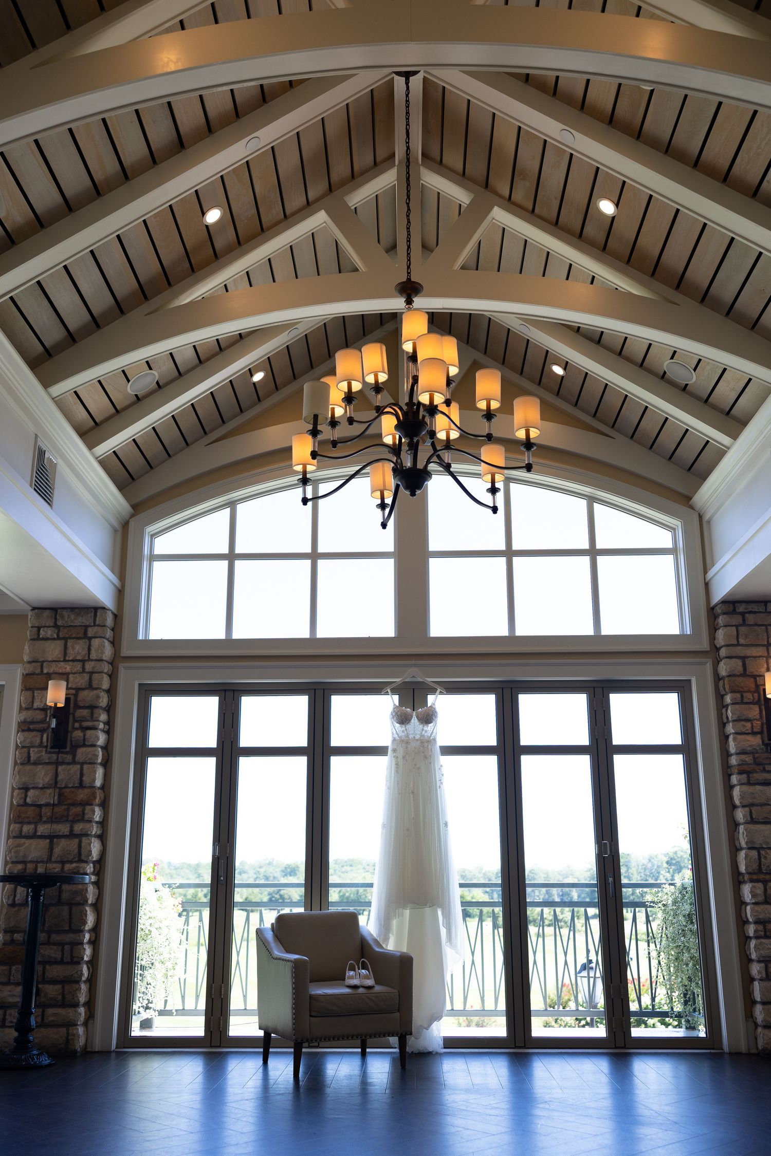 Wedding dress hanging from chandelier in elegant vaulted ceiling venue with large windows and stone walls.