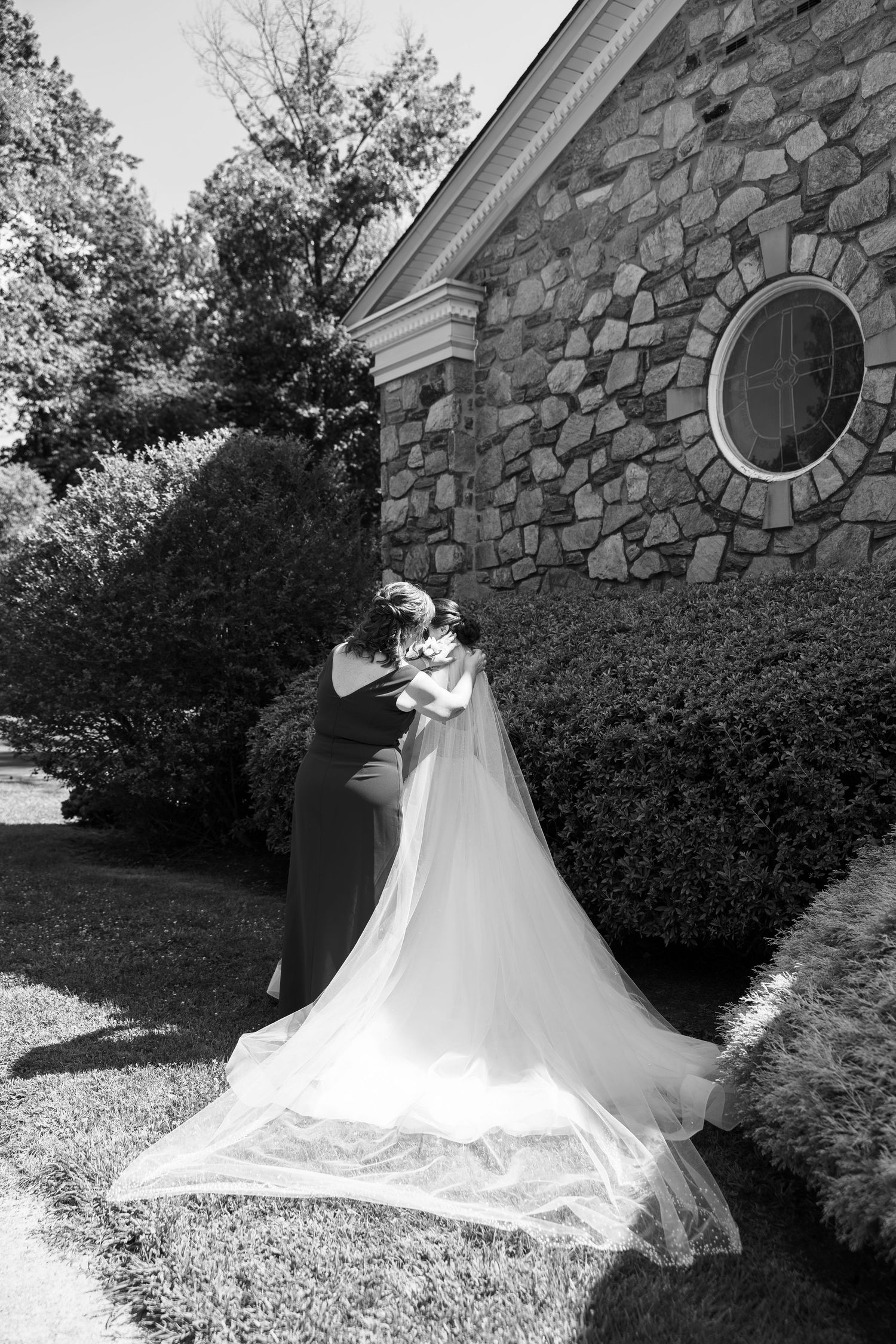 Wedding dress train spreads elegantly across stone building grounds in black and white. Mom helping with daughters veil.