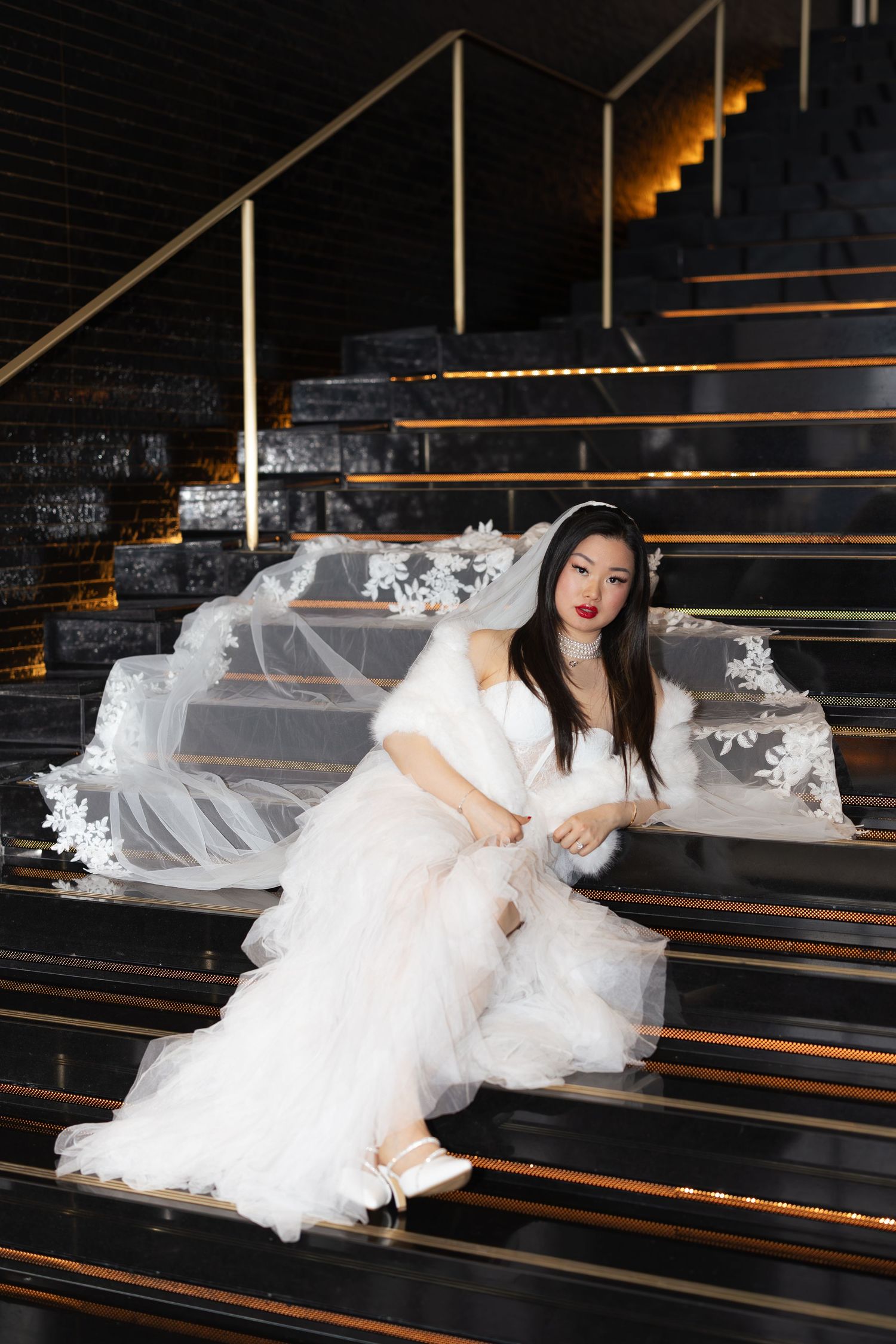 A bride in a white gown with fur trim sits elegantly on dark stairs with flowing tulle.