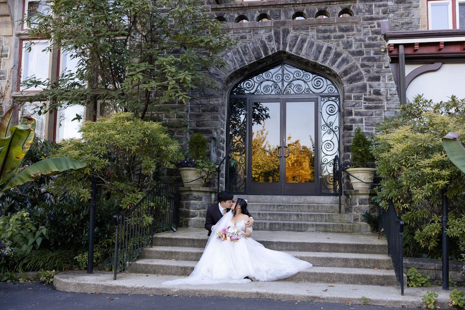 Wedding couple sitting by stone architecture with arched entryway surrounded by trees and landscaping.