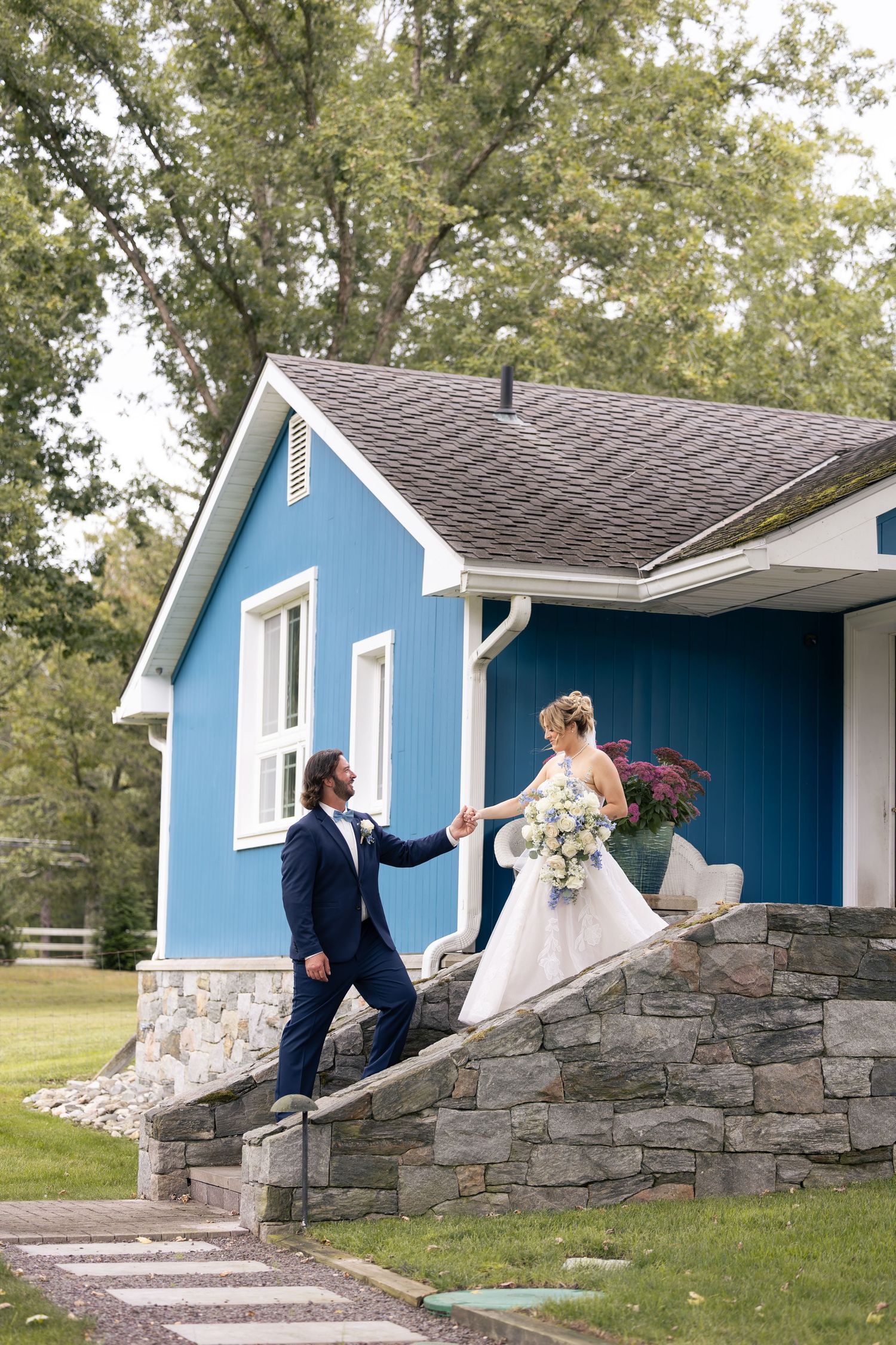 Bride and groom hold hands on stone steps outside a blue cottage surrounded by lush trees on their wedding day.
