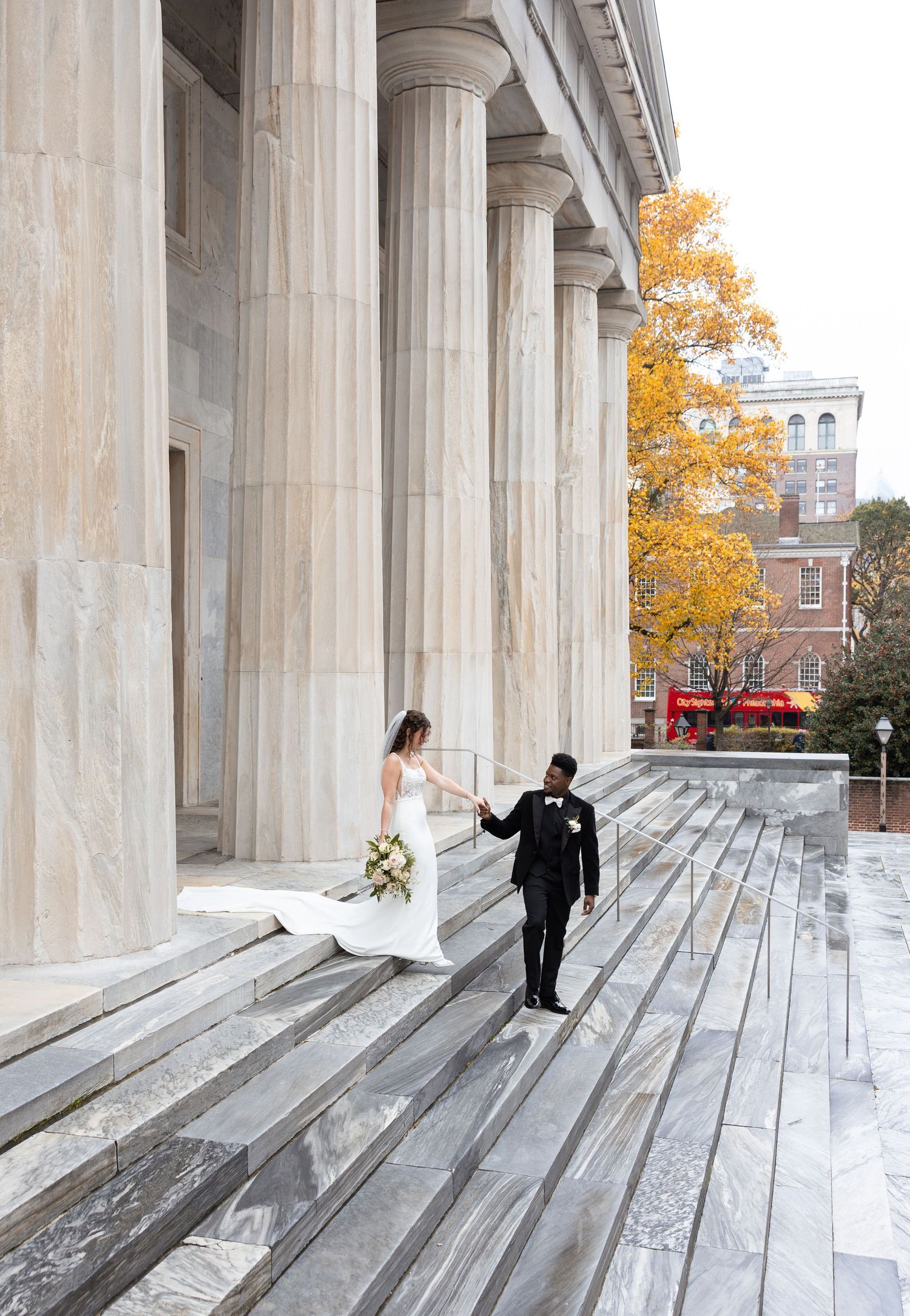A wedding couple descends grand marble steps with autumn foliage in the background.
