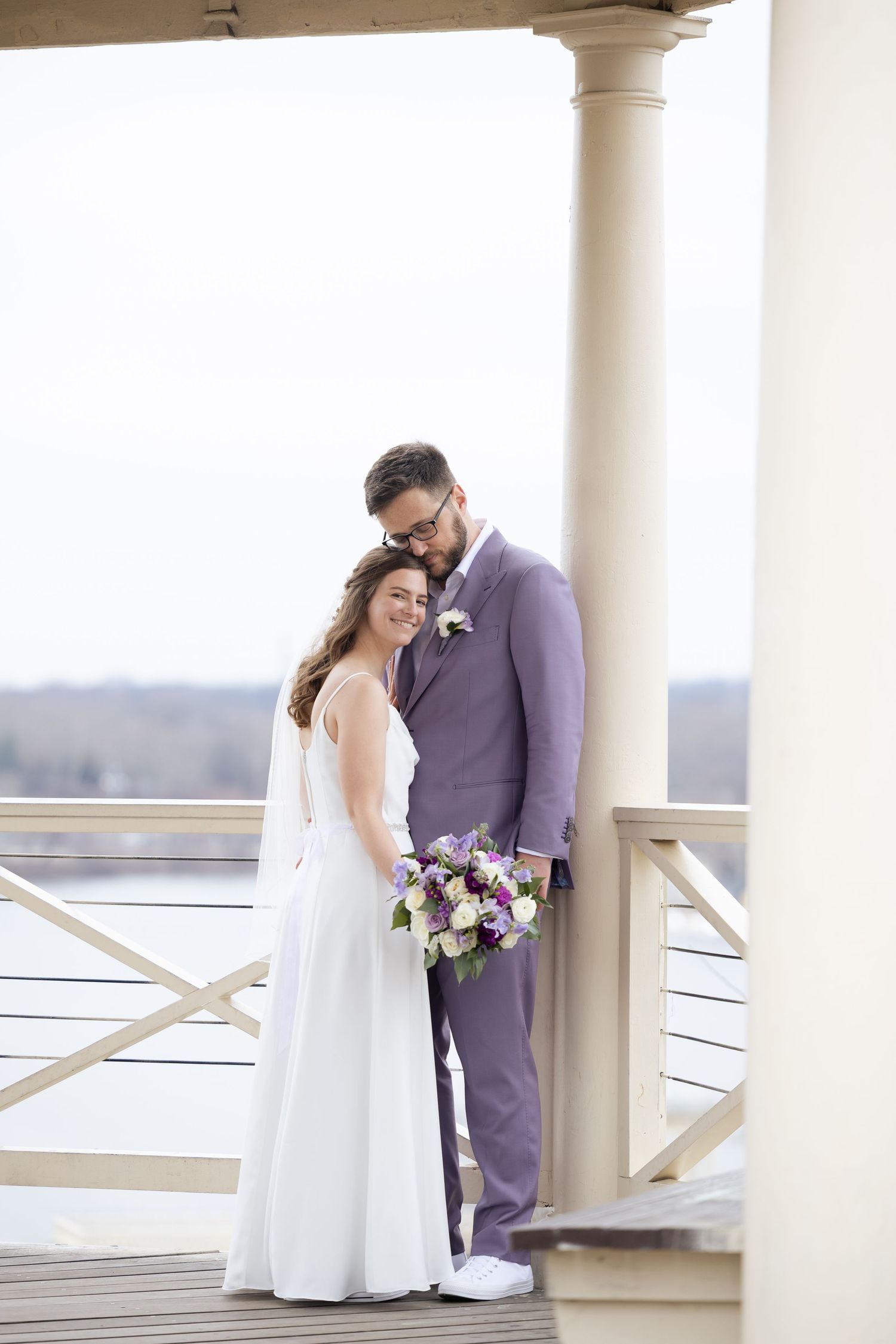Romantic wedding couple embracing on white columned gazebo with purple floral bouquet.