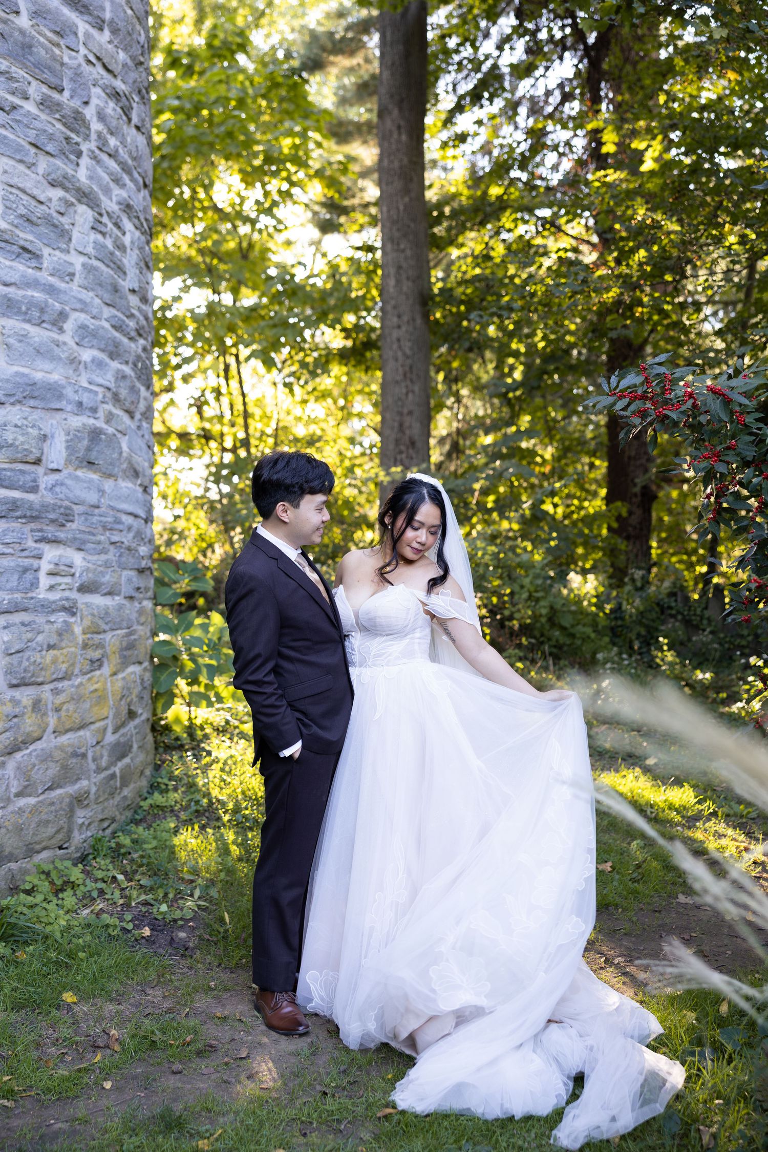 Wedding couple poses by stone wall in sunlit garden setting.