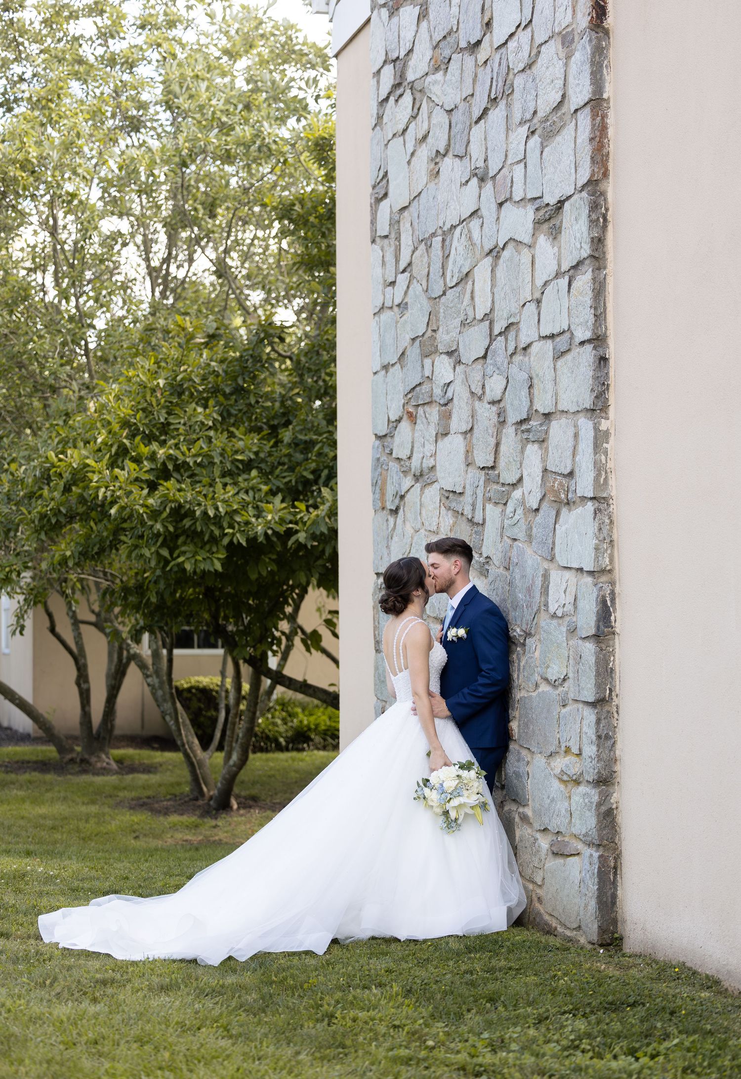 Wedding couple poses against textured stone wall with lush greenery in background.