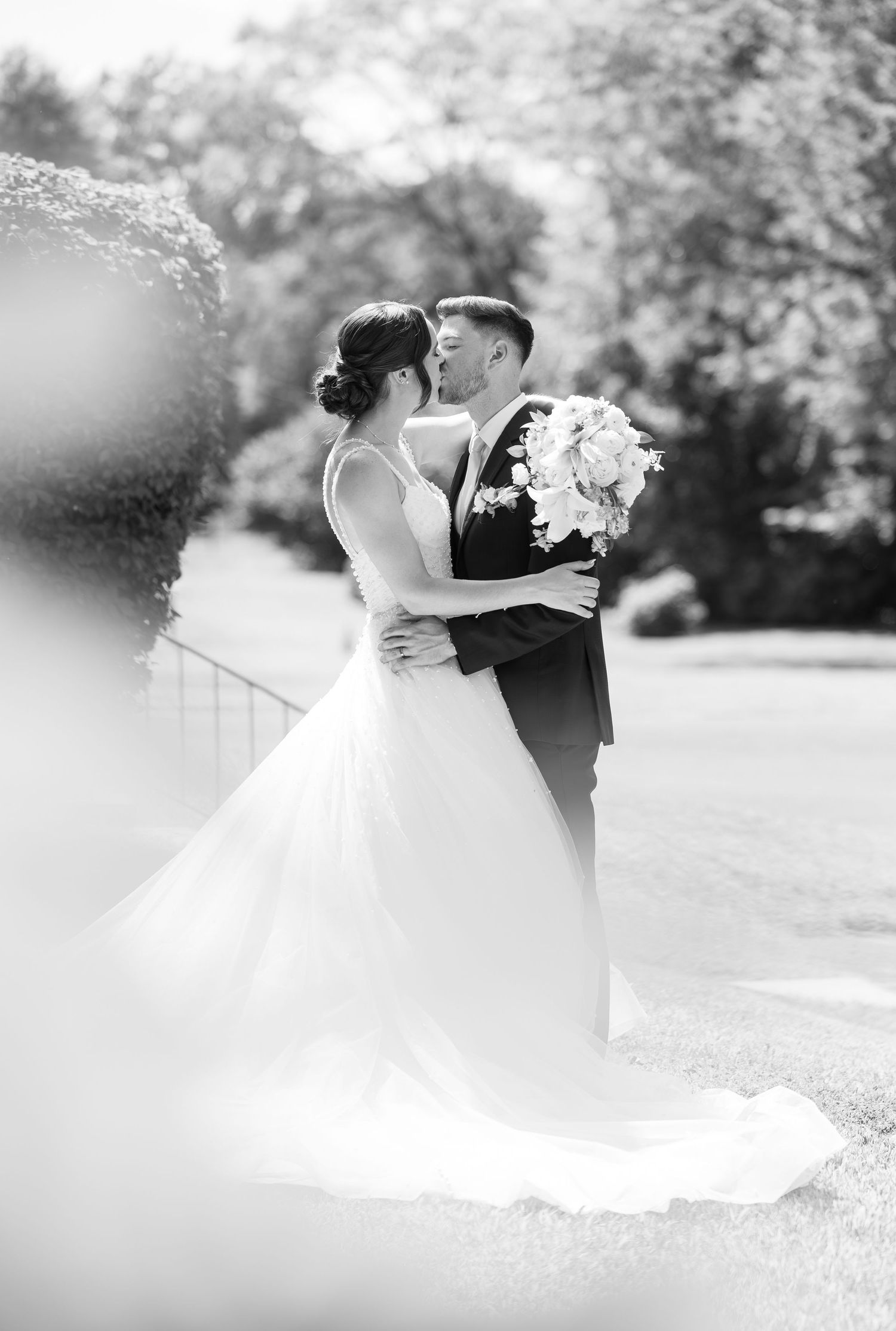 A black and white portrait of newlyweds embracing outdoors with soft focus foreground elements.