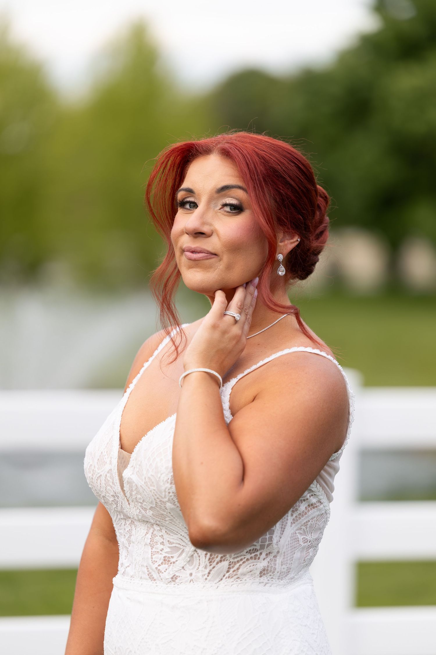 Red-haired bride in white lace gown poses gracefully outdoors near white fence, touching her neck and smiling softly.