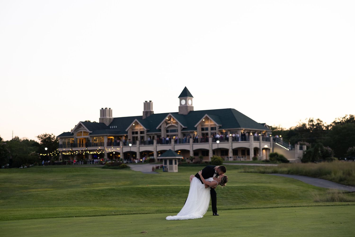 Bride and groom share romantic dip kiss on golf course lawn with elegant wedding venue clubhouse in background at dusk.