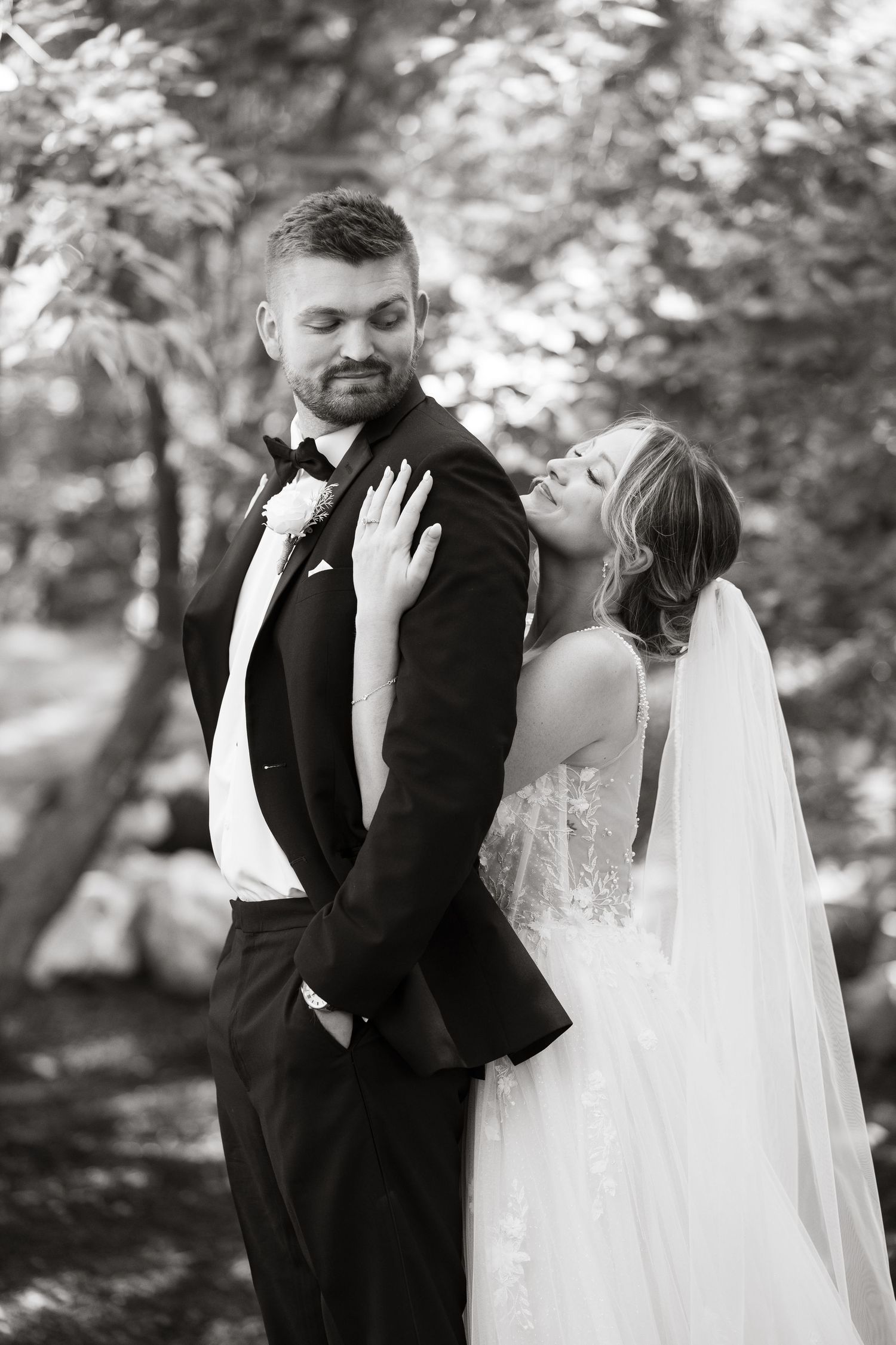 Black and white photo of a bride in lace gown leaning on groom in tuxedo outdoors among trees.