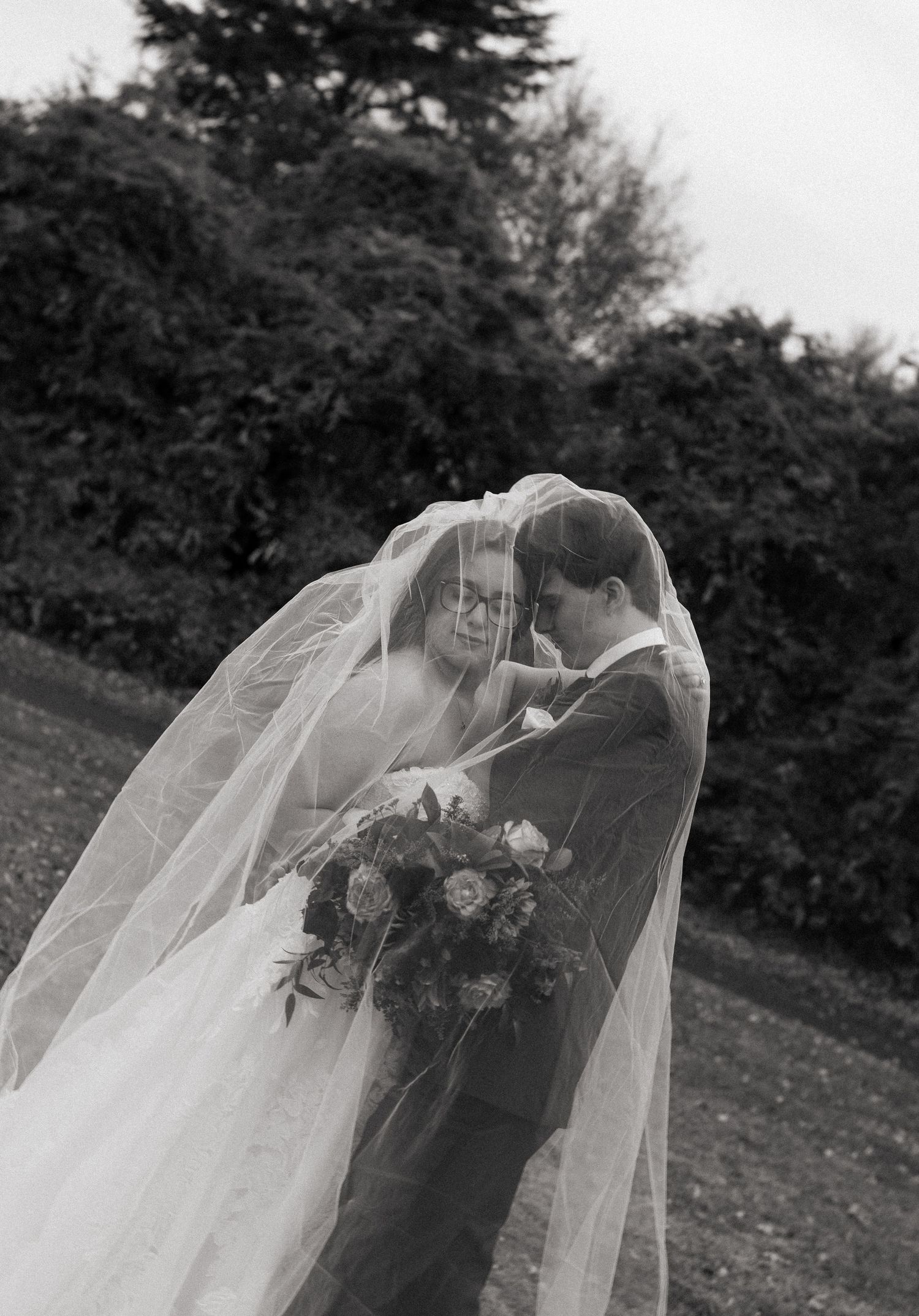 Black and white photo of bride and groom sharing an intimate moment under a flowing veil outdoors.