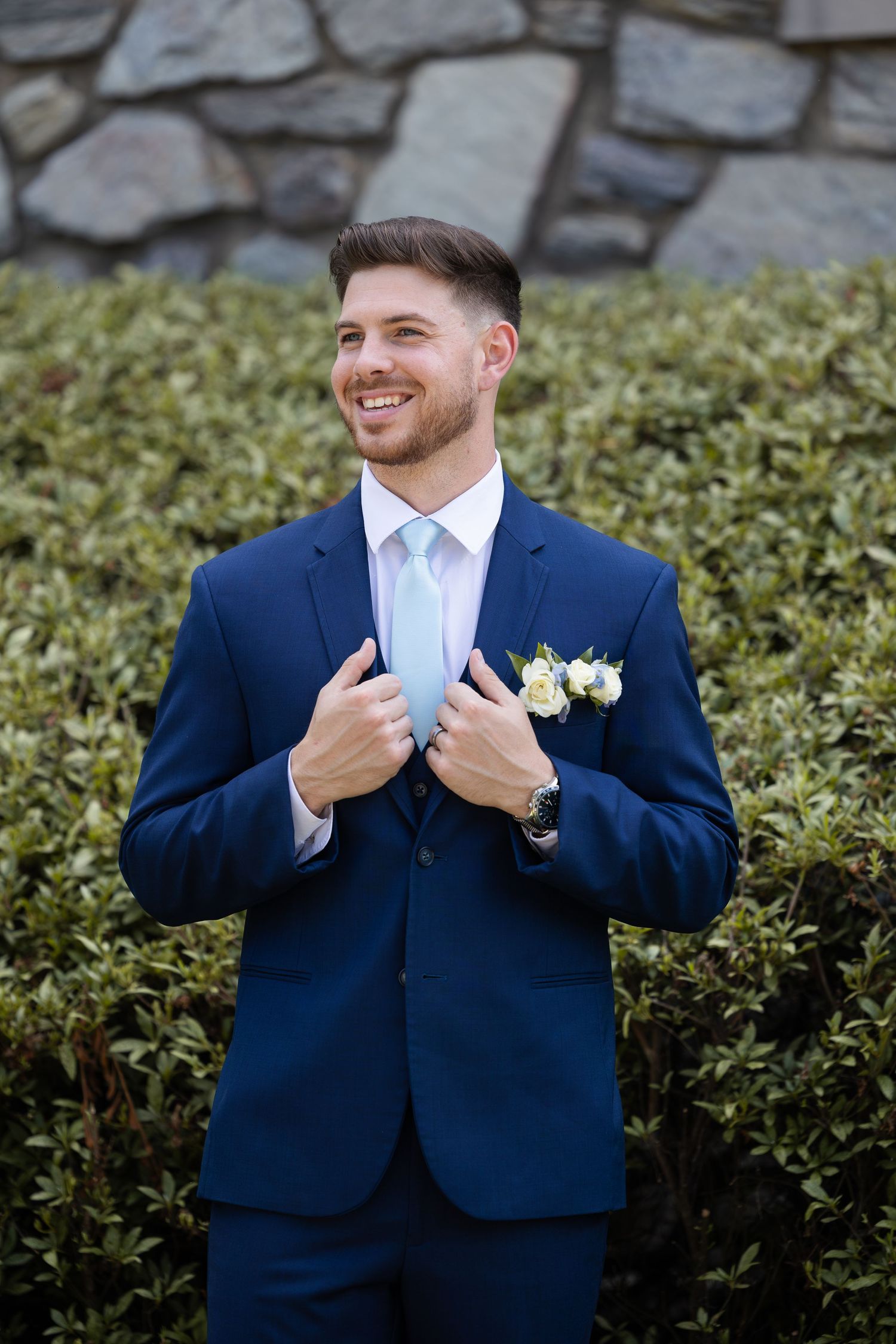 Groom in navy suit adjusts his tie while smiling against stone wall and greenery backdrop.