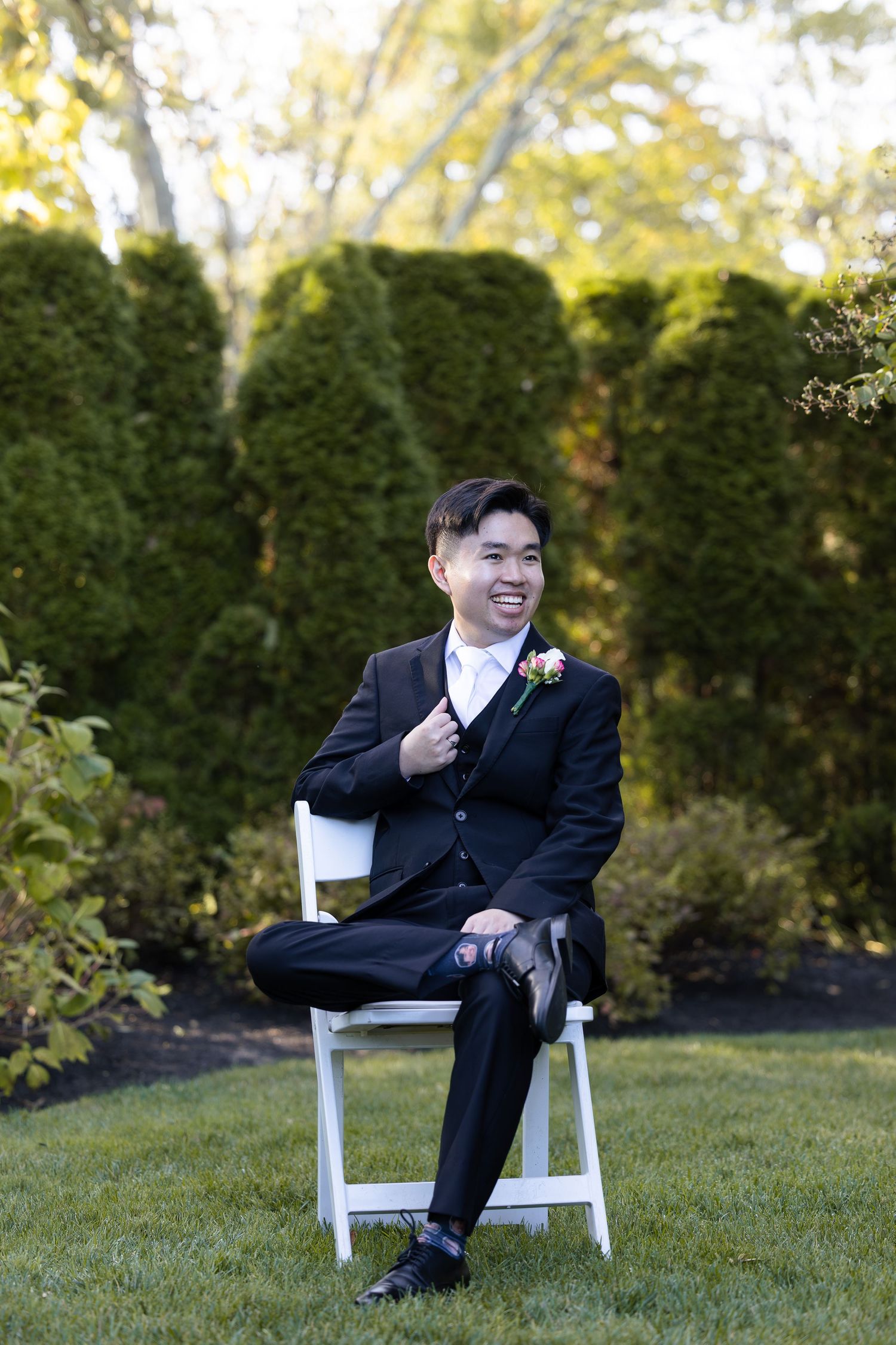 Young man in black tuxedo with boutonniere sits on white chair in garden, smiling confidently at outdoor wedding.