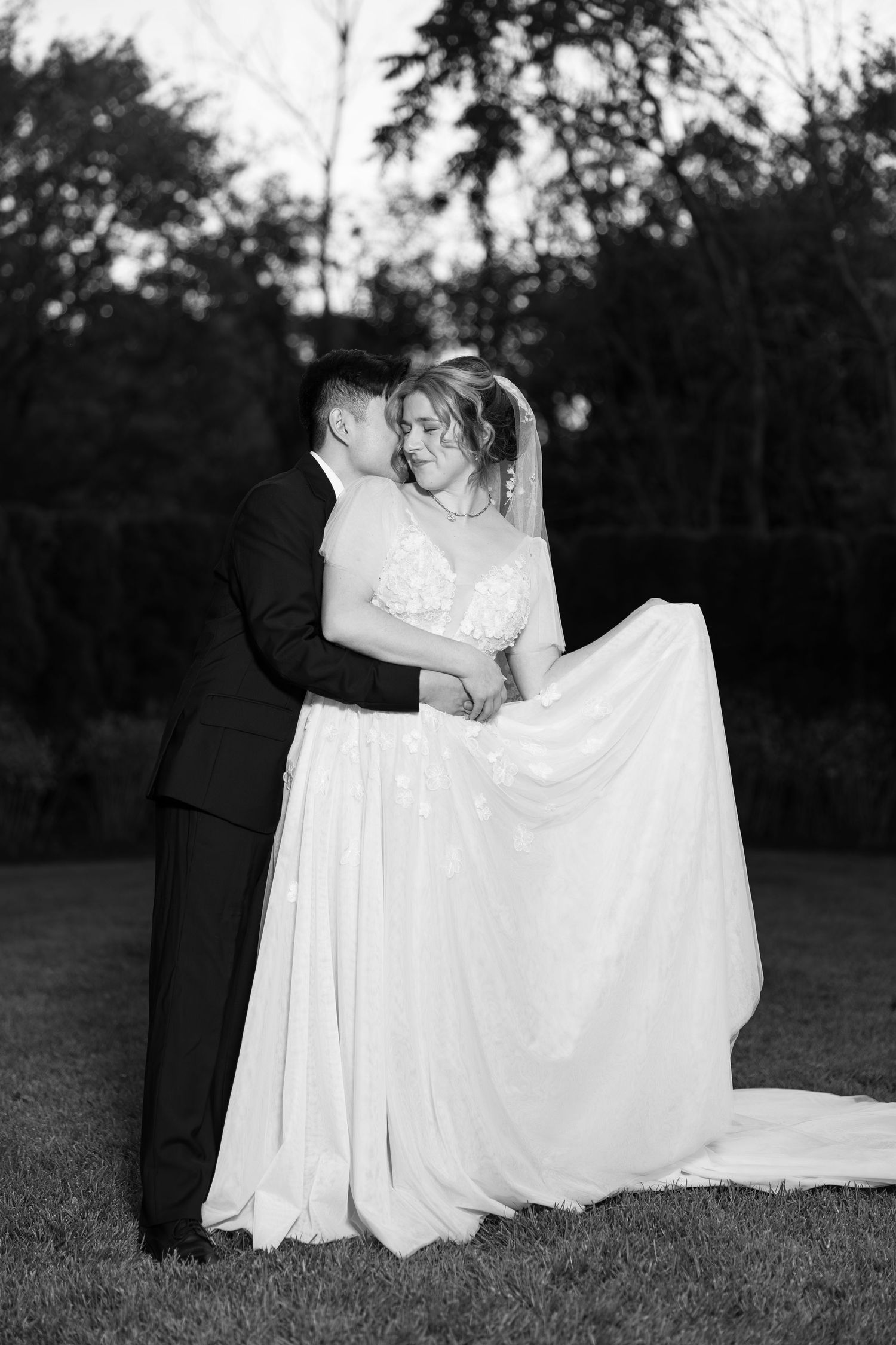 Black and white photo of a bride and groom embracing outdoors, bride in flowing gown with veil, groom in suit.