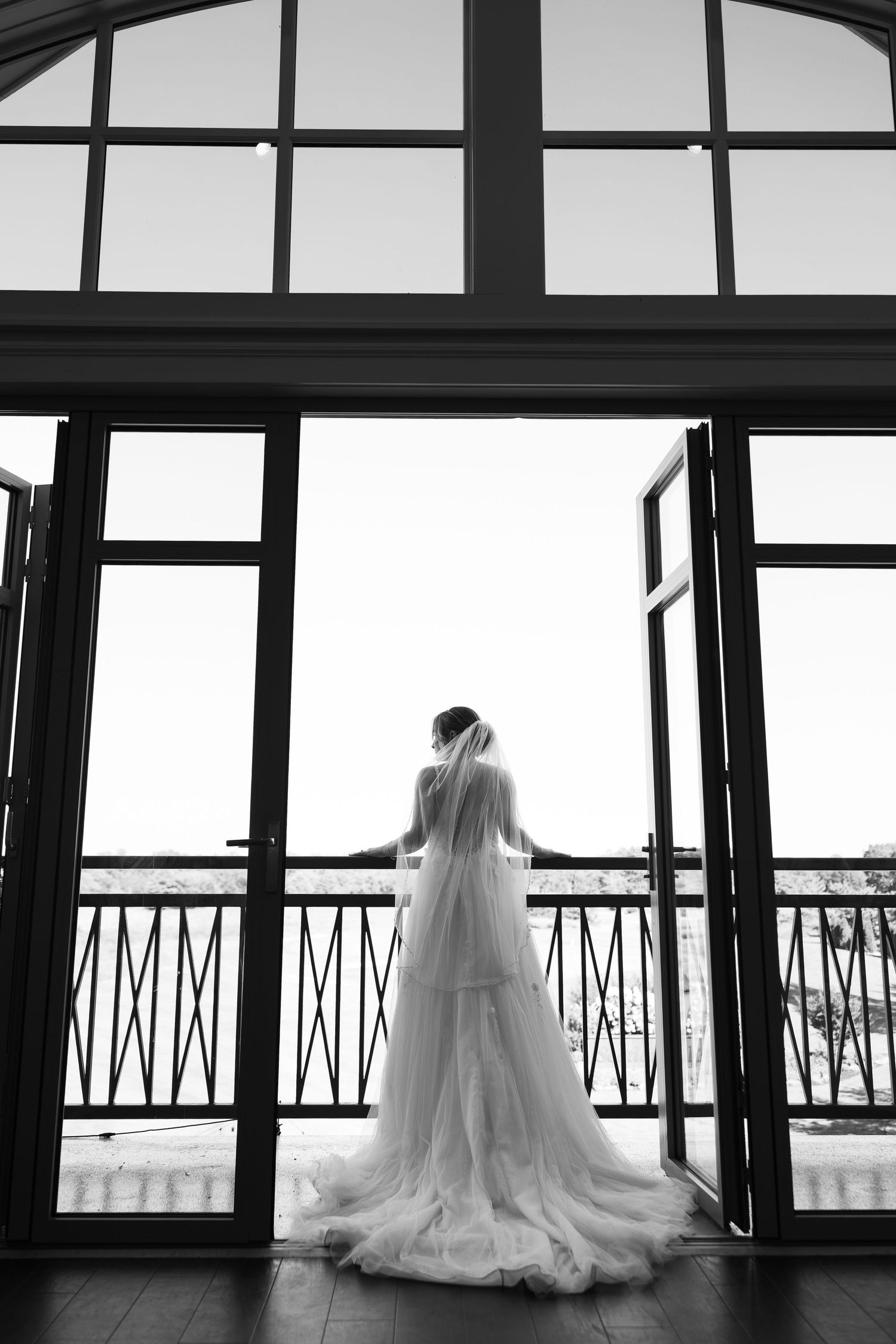 Silhouette of bride in flowing dress standing at elegant glass doors overlooking balcony.